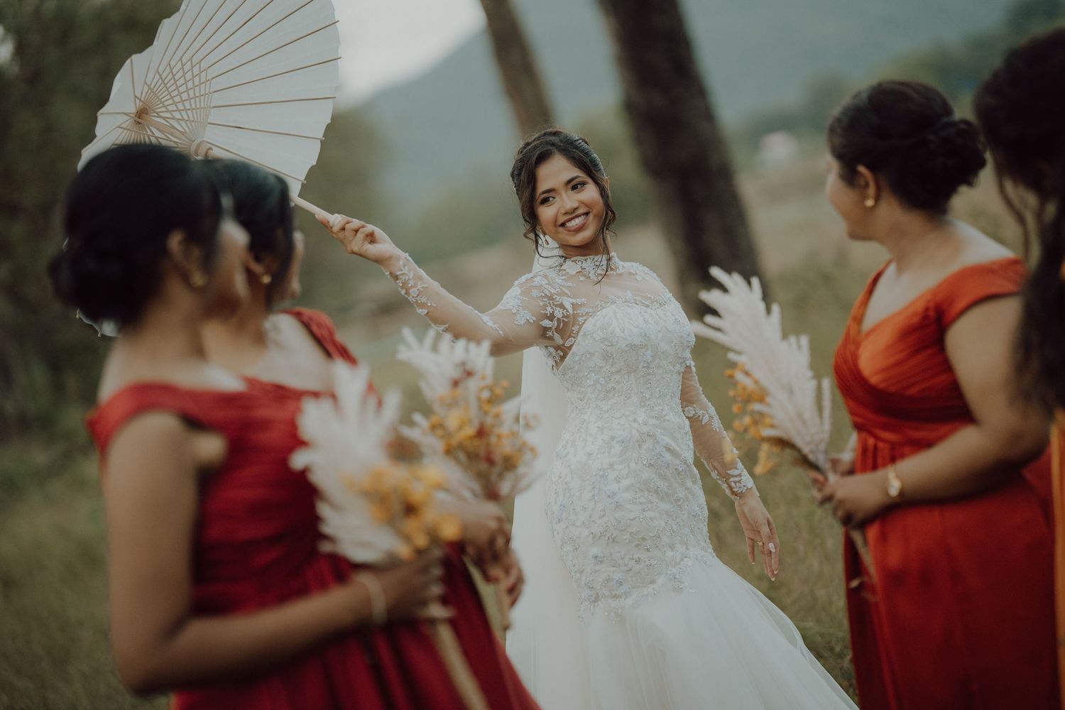 Joyful bride in a lace wedding dress holding a white fan laughs with bridesmaids in red dresses outdoors.