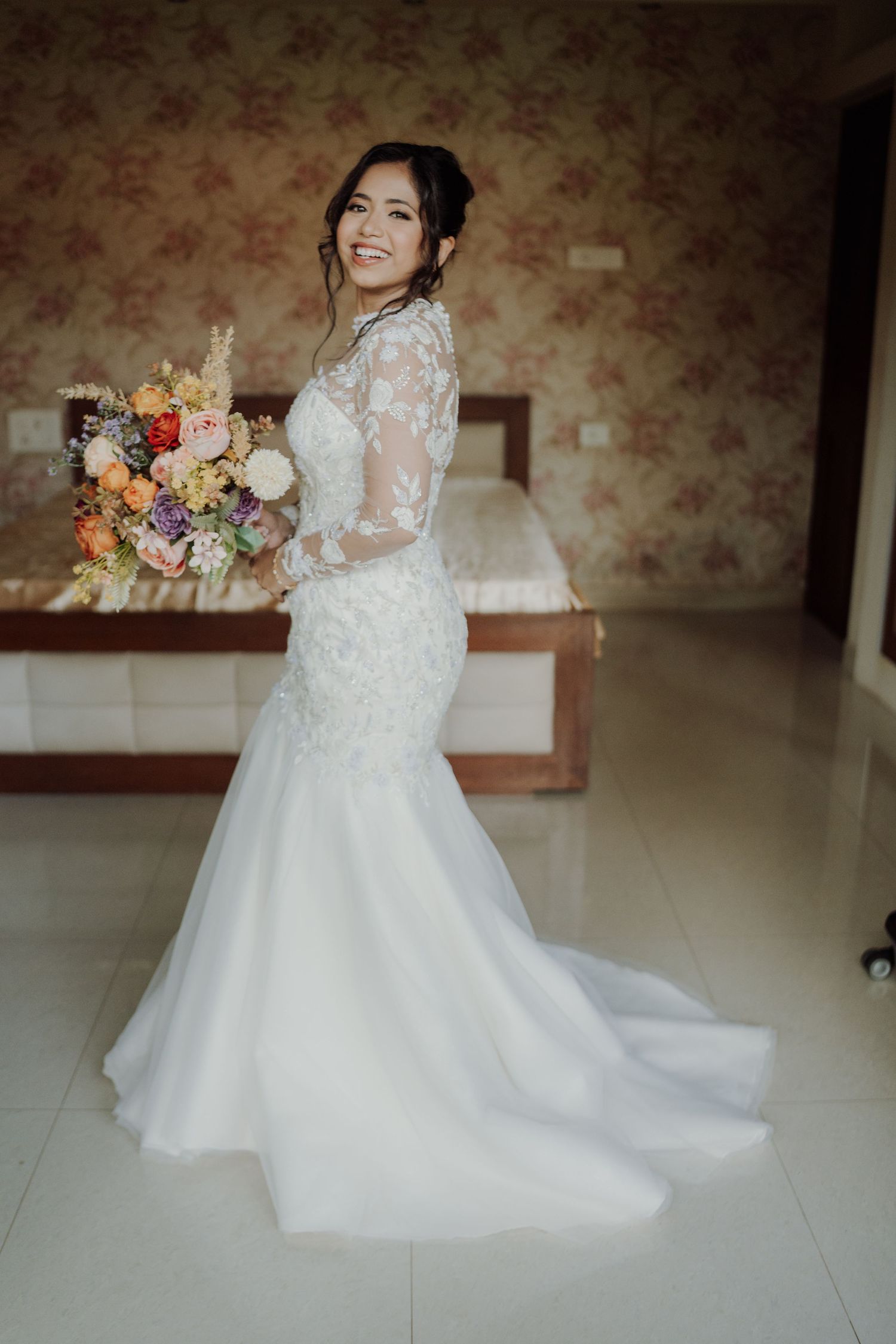 Smiling bride in a white lace mermaid wedding gown holding a colorful bouquet in a bright bedroom.