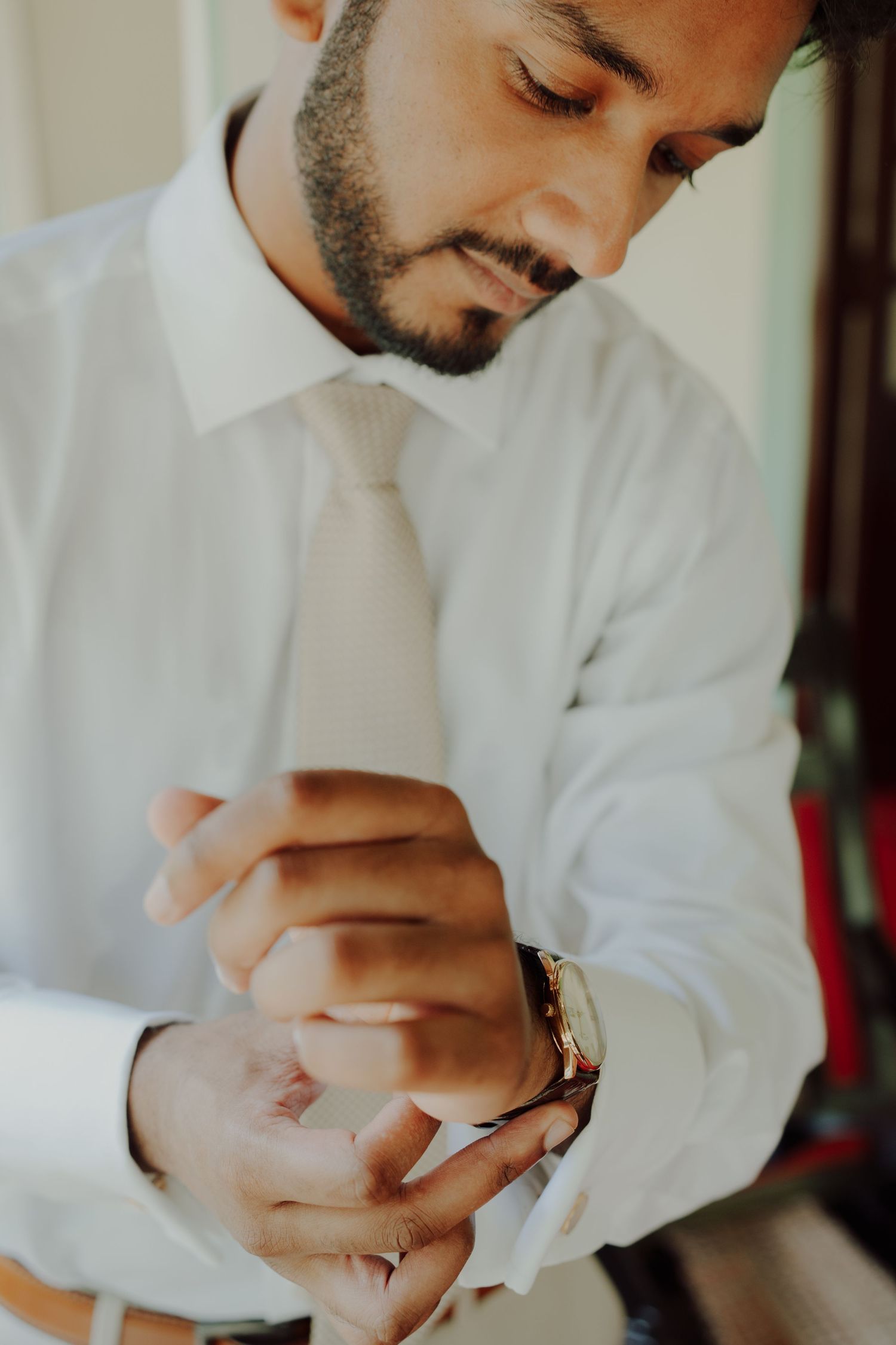 Groom in a white dress shirt and beige tie adjusts his wristwatch and cuff while getting ready on his wedding day.
