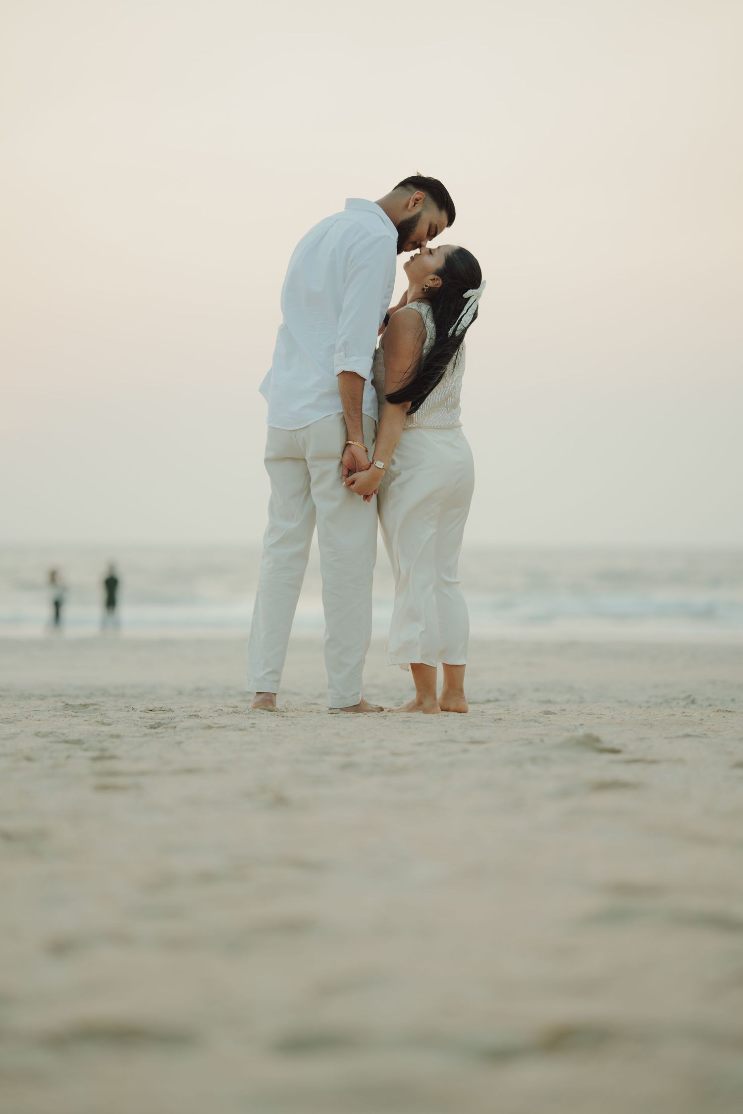 Couple in white outfits sharing a romantic kiss on a sandy beach at dusk with ocean waves in the background.