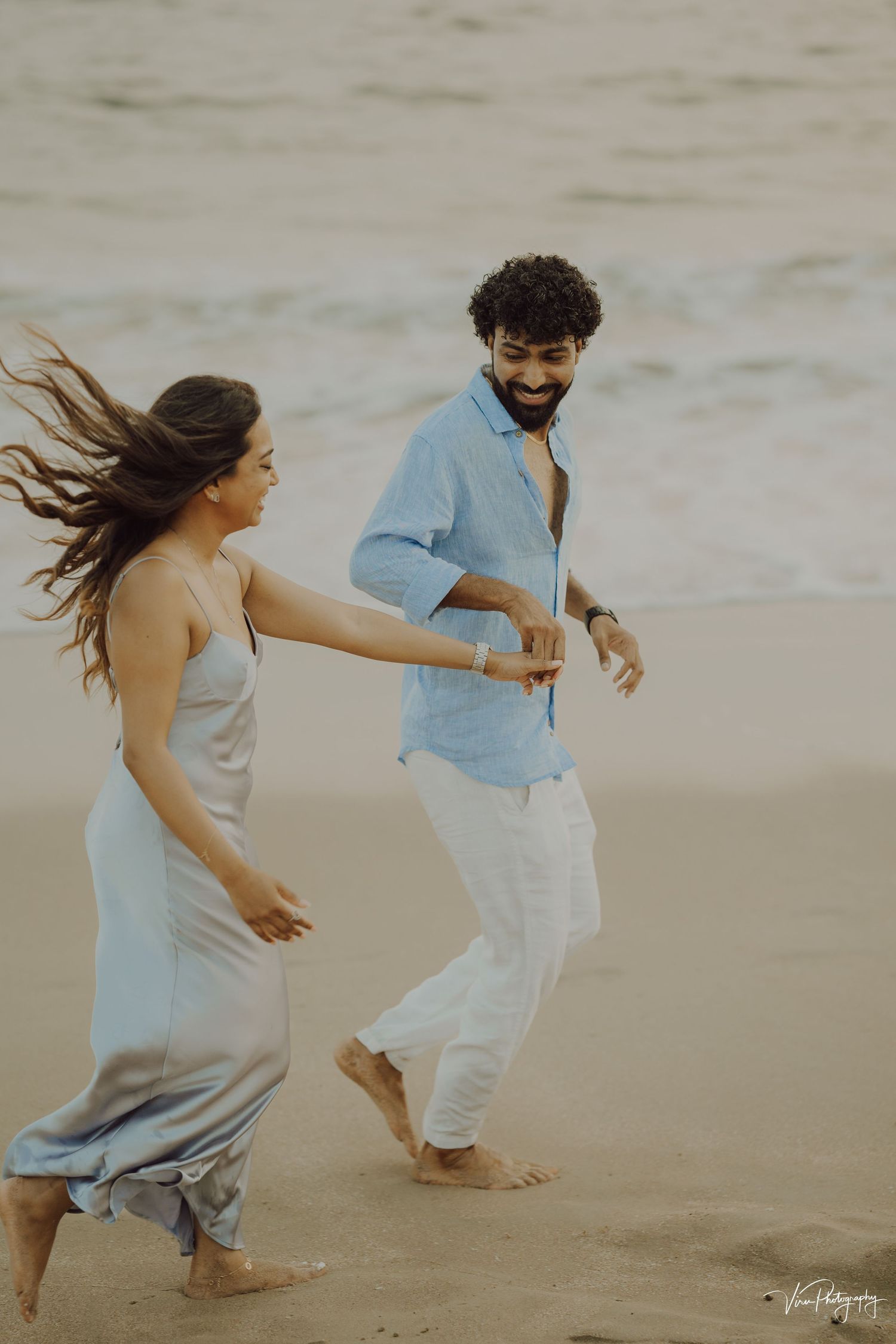 A couple dances joyfully on the beach with windswept hair, wearing light colored casual outfits in a candid moment.
