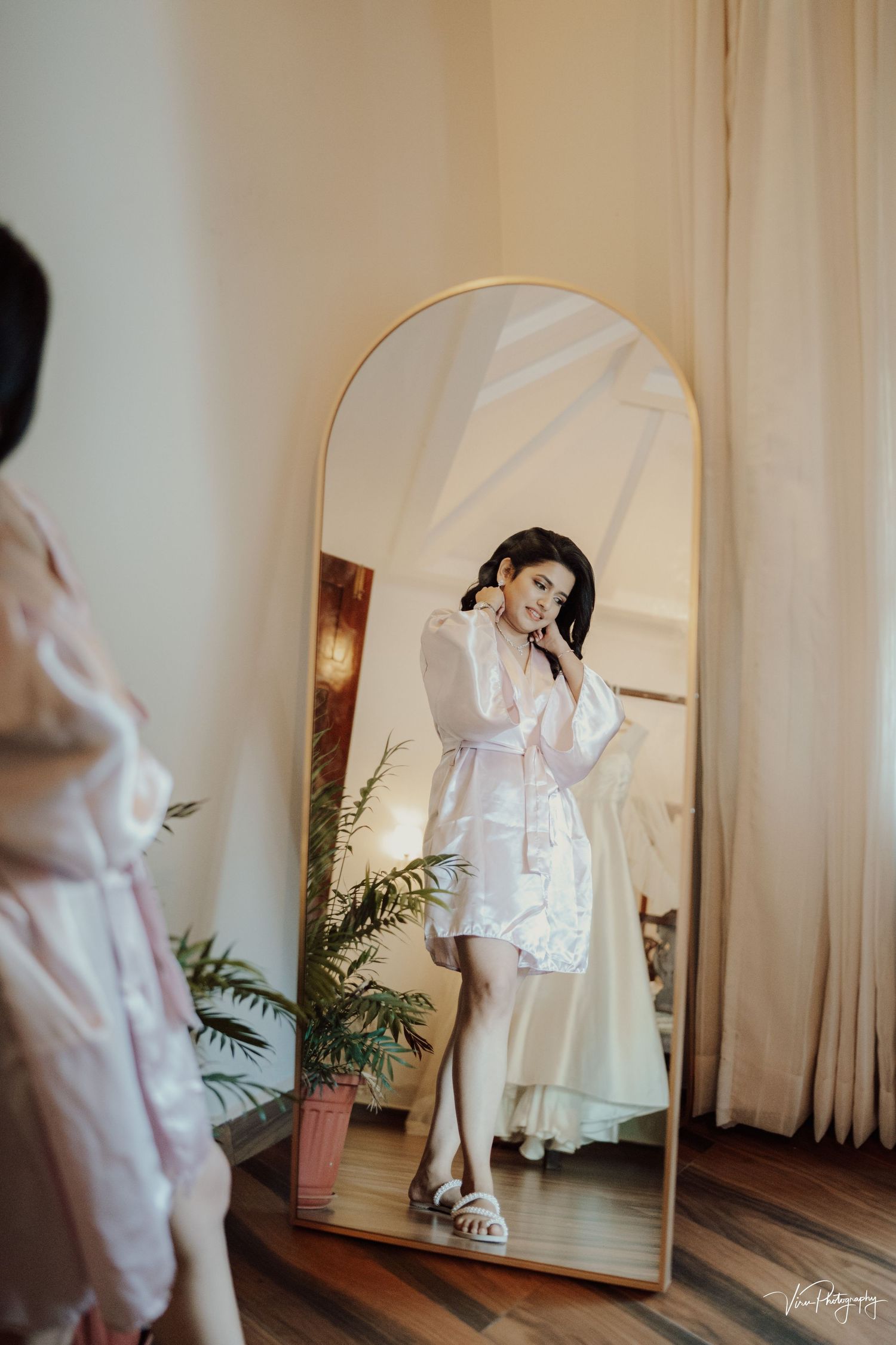 A silhouetted figure in a white robe stands before an arched floor mirror with houseplants and curtains in a bright room.