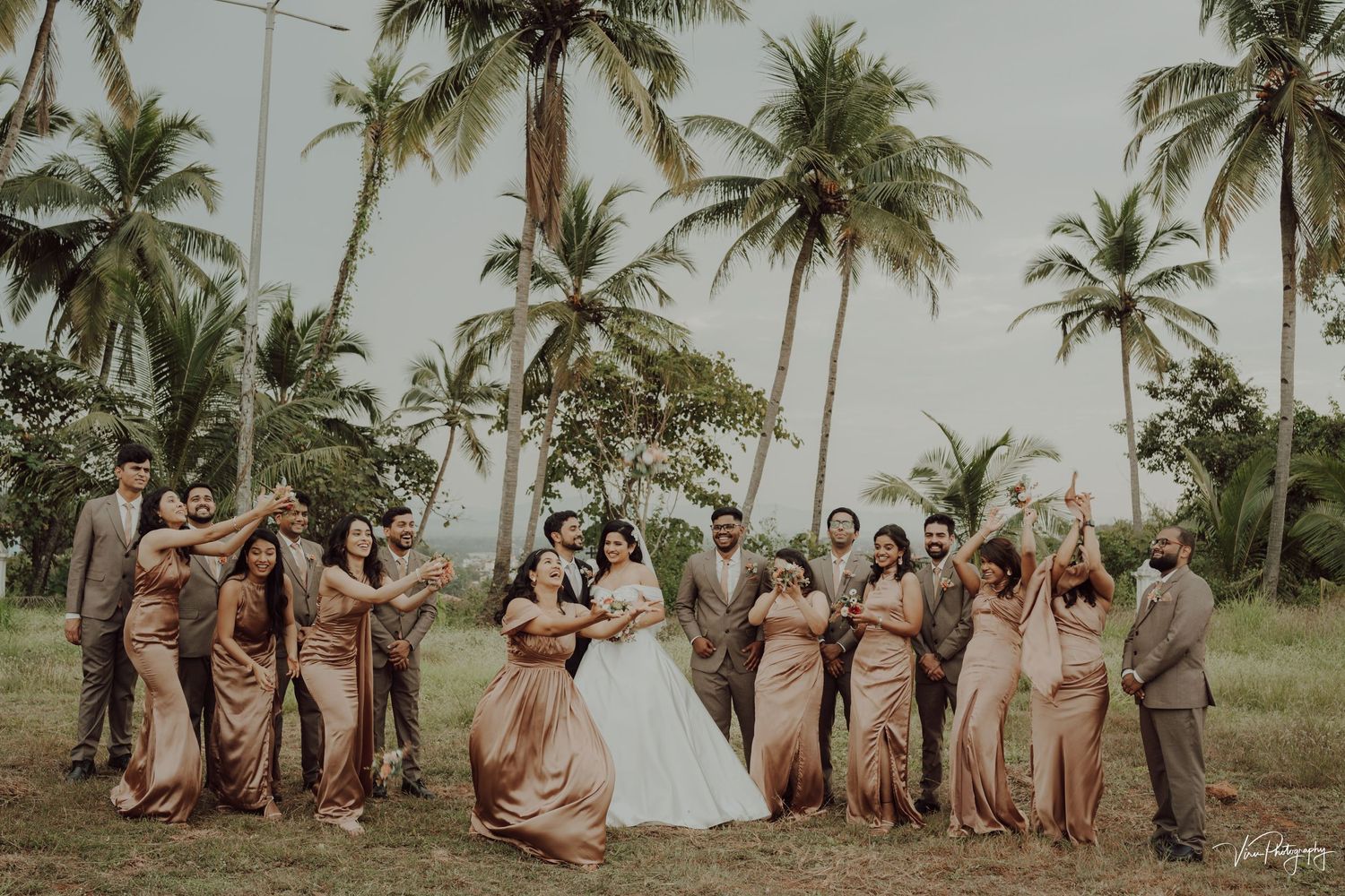 Wedding party wearing blush pink and brown attire pose together under tall palm trees in a tropical setting.