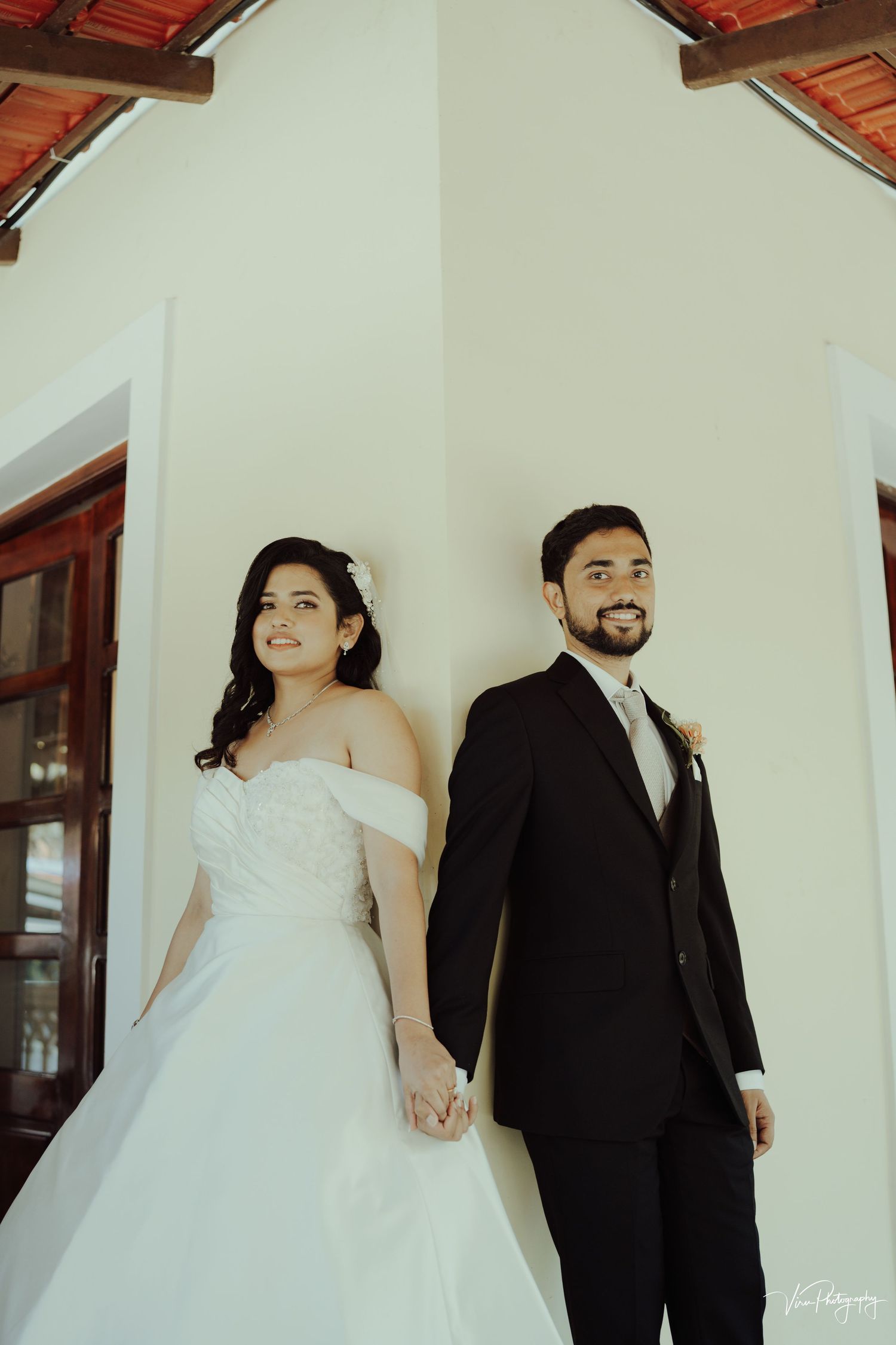 Newlyweds hold hands against white wall beneath terracotta roof in romantic wedding portrait.