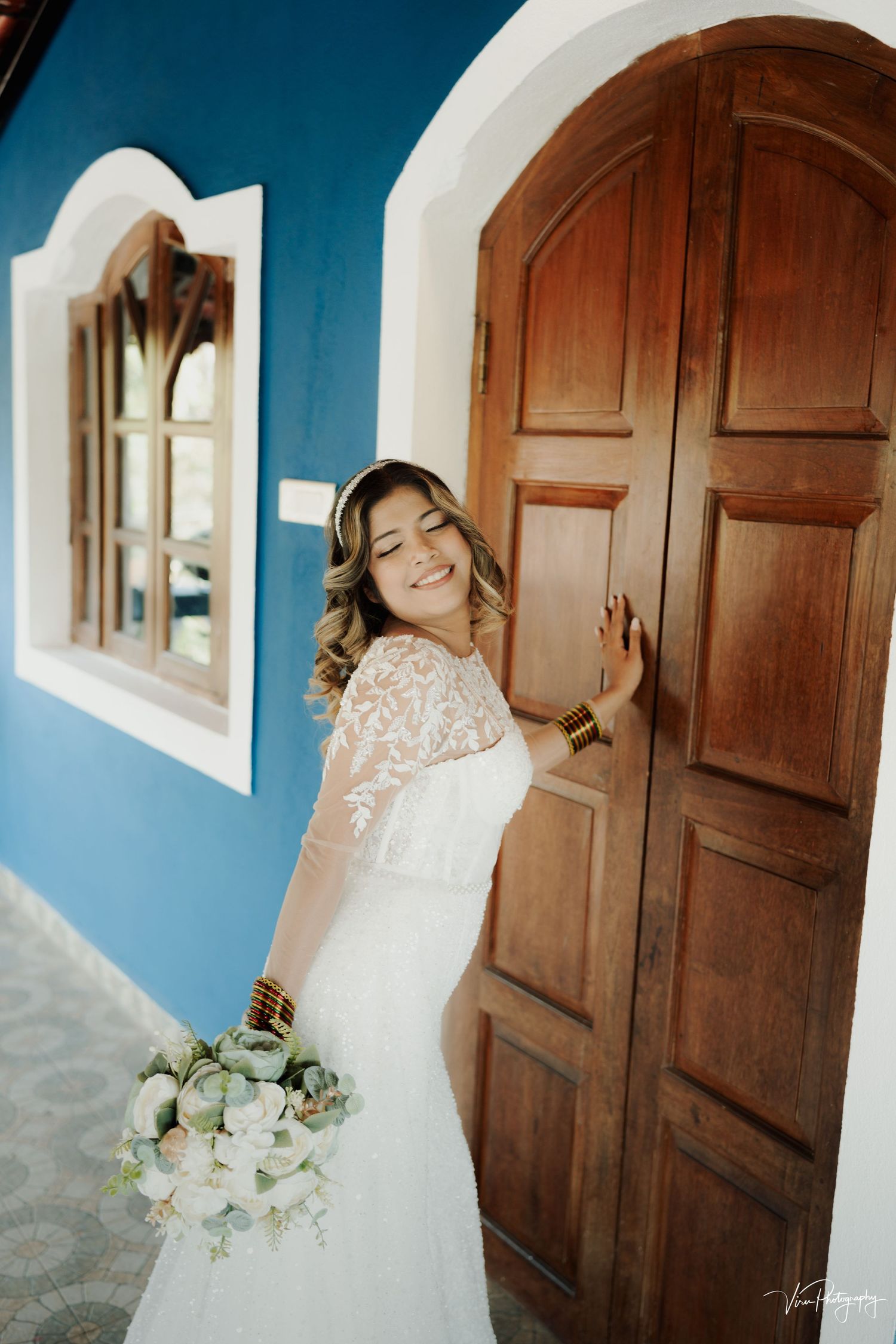 A bride in a white lace gown holds a bouquet of white flowers while posing by a wooden door against bright blue walls.