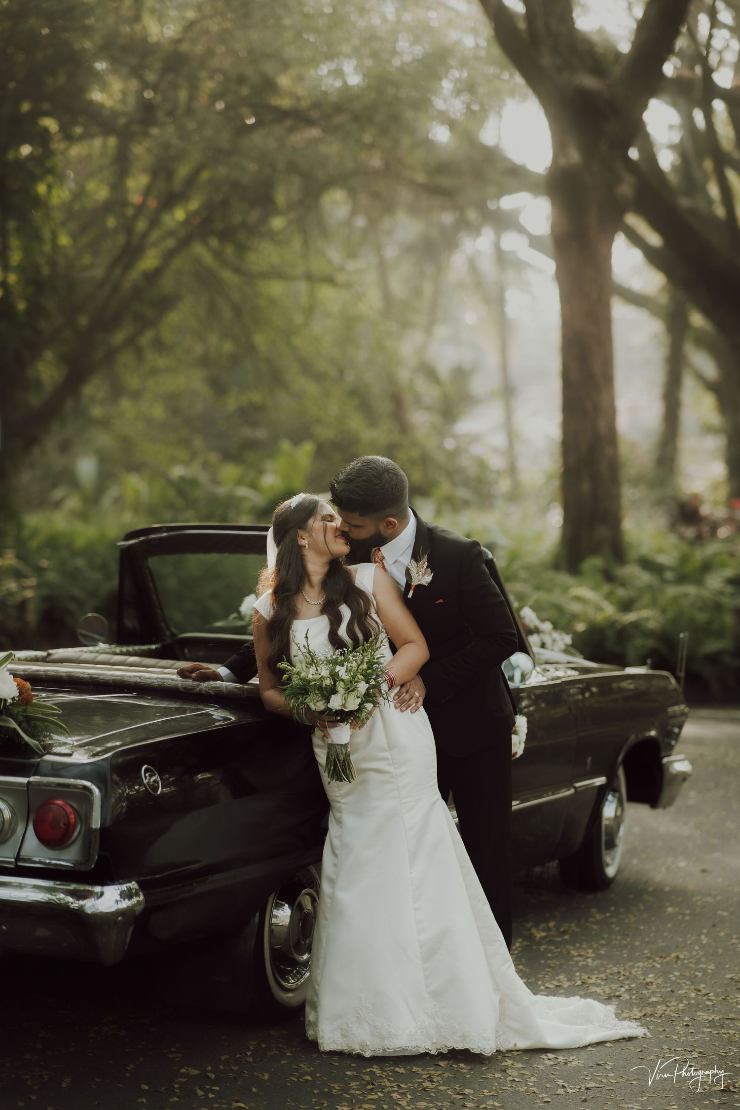 A romantic wedding scene with a couple by a classic black car on a misty forest road.