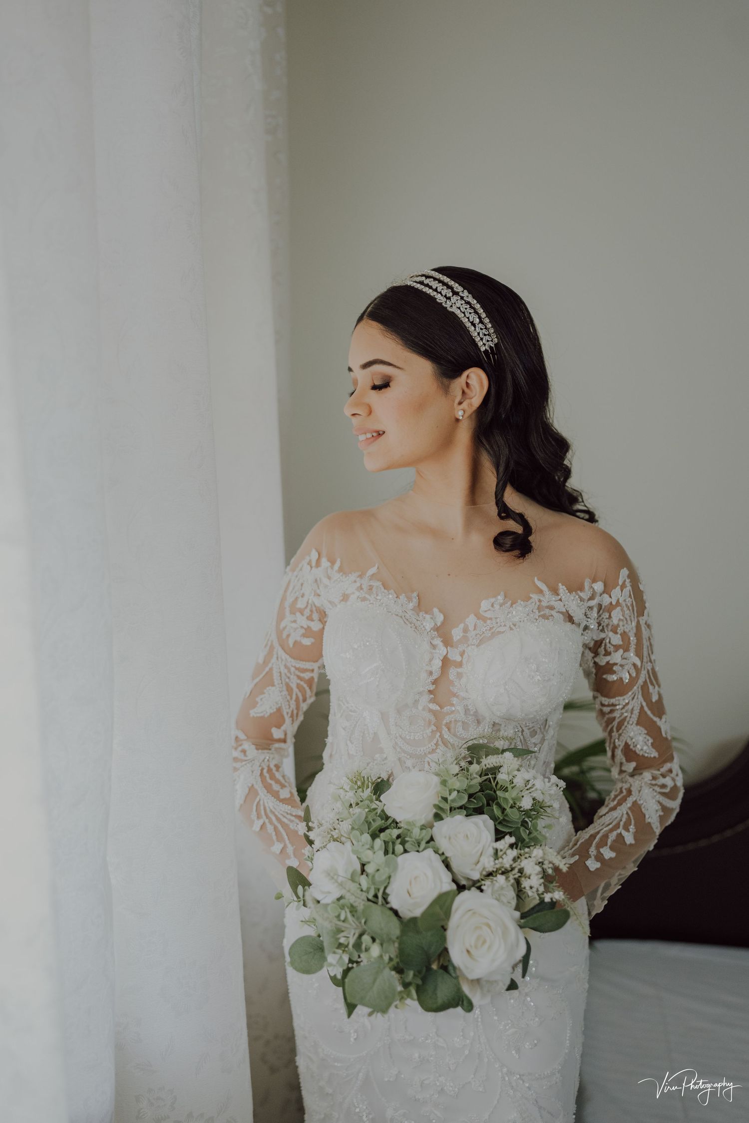 Beautiful bridal portrait showing a white lace long-sleeve wedding gown and elegant crystal headpiece with white rose bouquet.
