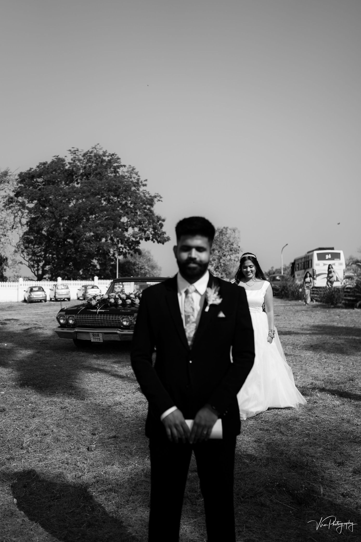 Black and white wedding photo showing bride and groom in an outdoor setting with trees and parked cars in background.