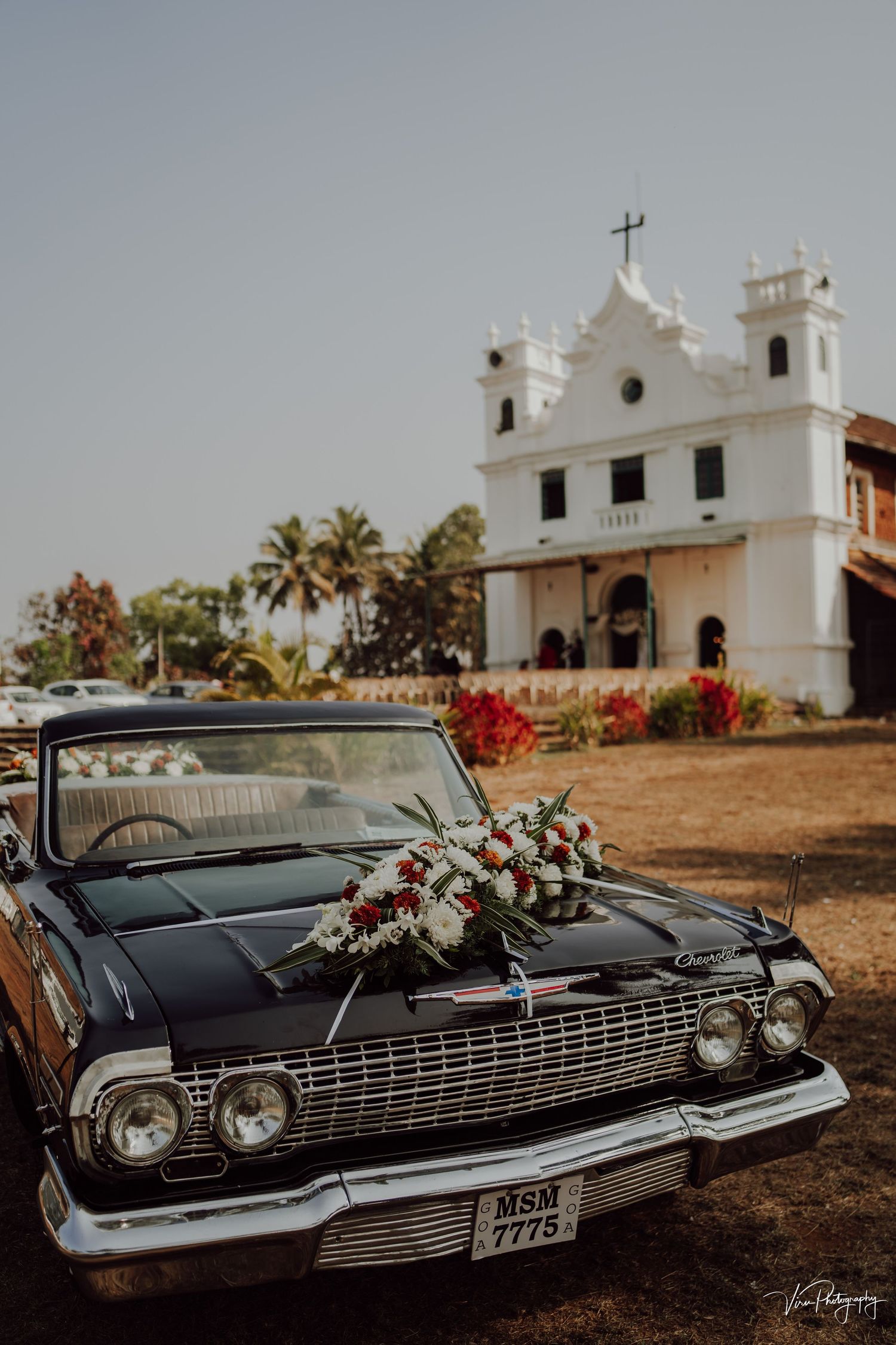 Classic black vintage car decorated with flowers parked in front of a white colonial church with palm trees in Goa.
