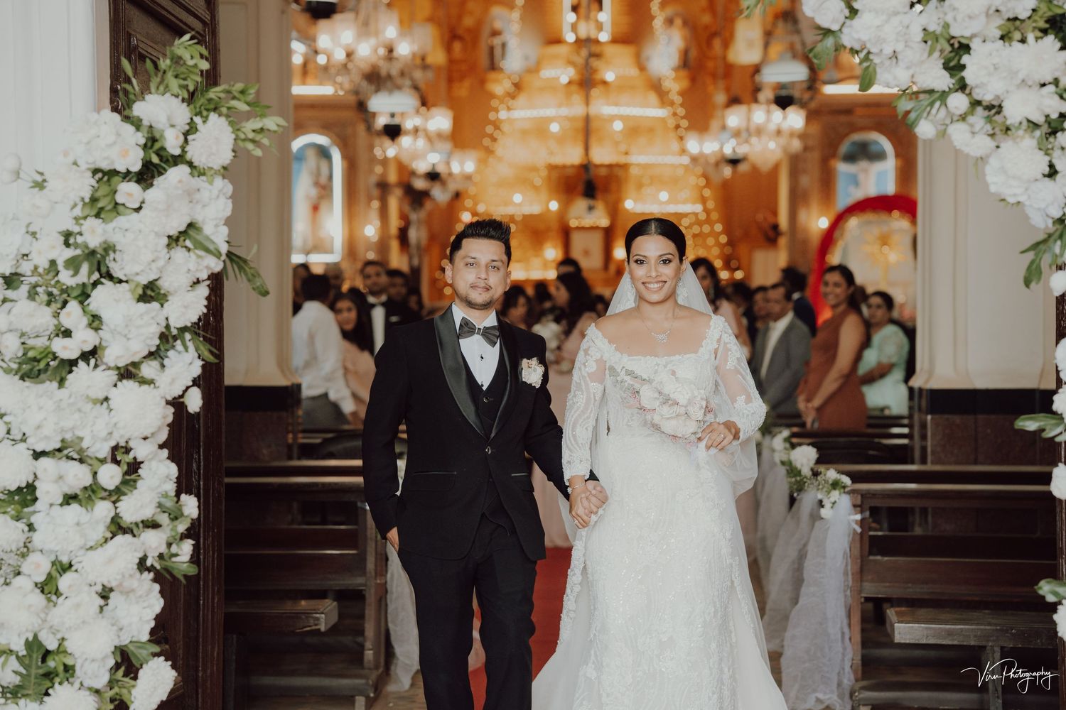 A wedding ceremony in a beautifully decorated church with white floral arches and rows of wooden pews.