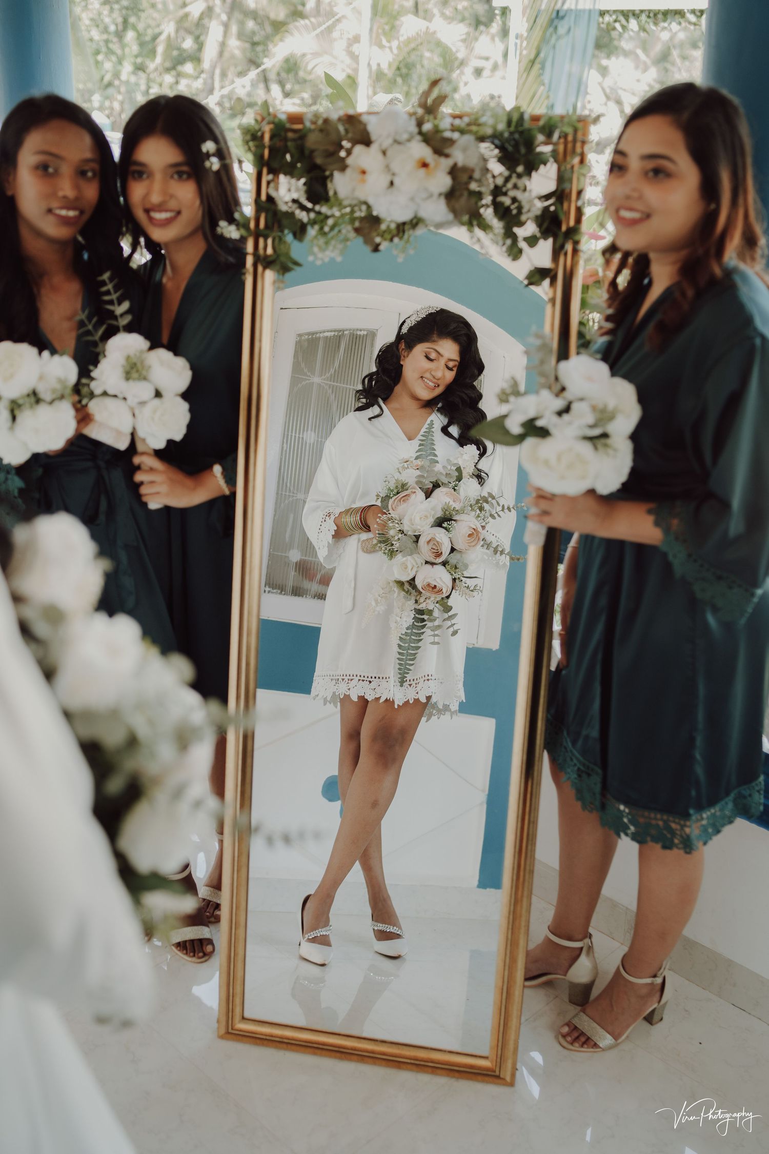 A bridal photo shoot scene with bridesmaids holding white bouquets and a gold-framed mirror reflection in a bright setting.