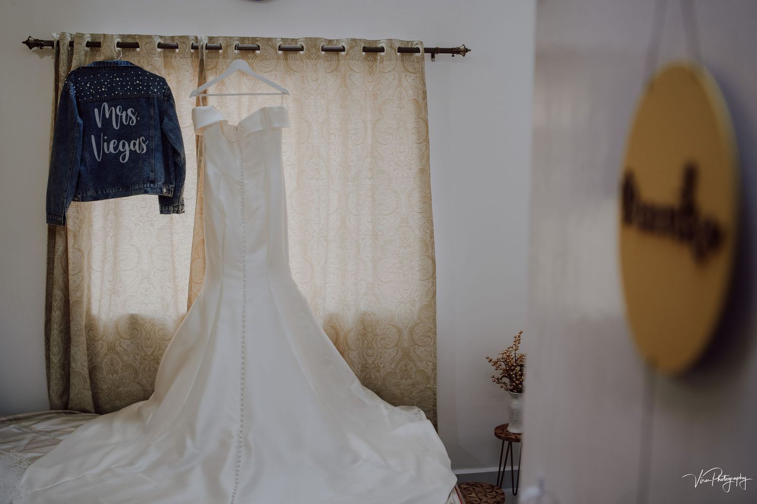 A white wedding dress hangs elegantly beside a dark blue denim jacket against cream curtains in a softly lit room.