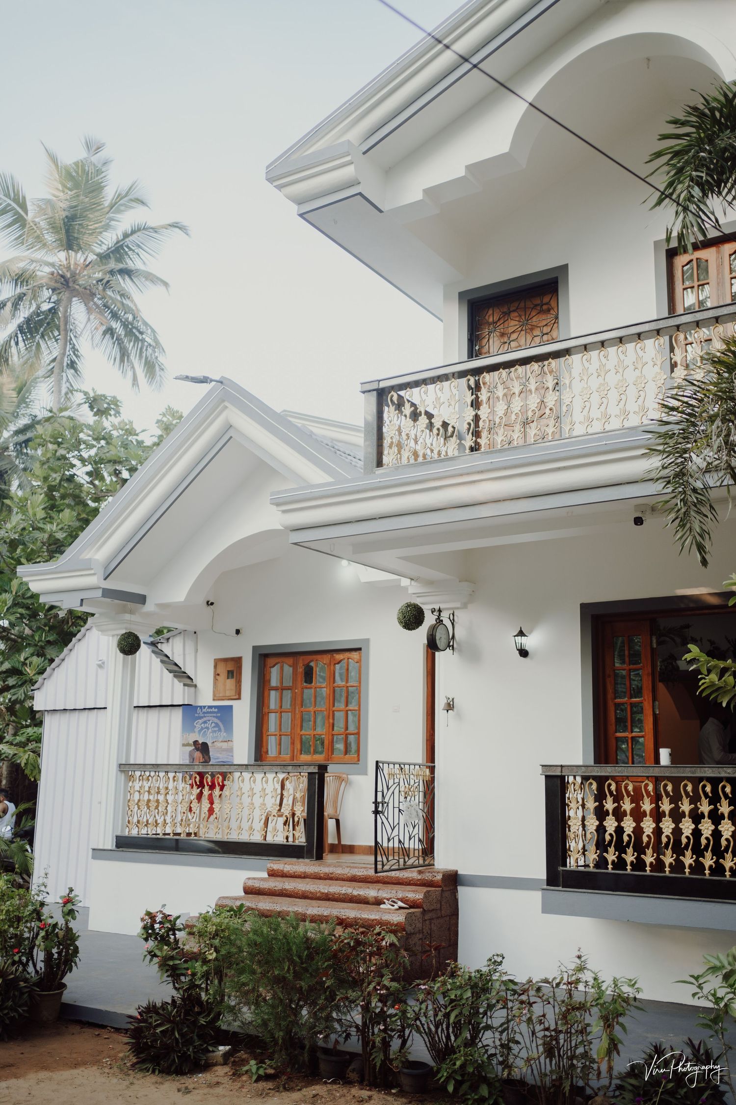 White two-story tropical house with wooden doors, ornate balconies, and palm trees in a peaceful residential setting.