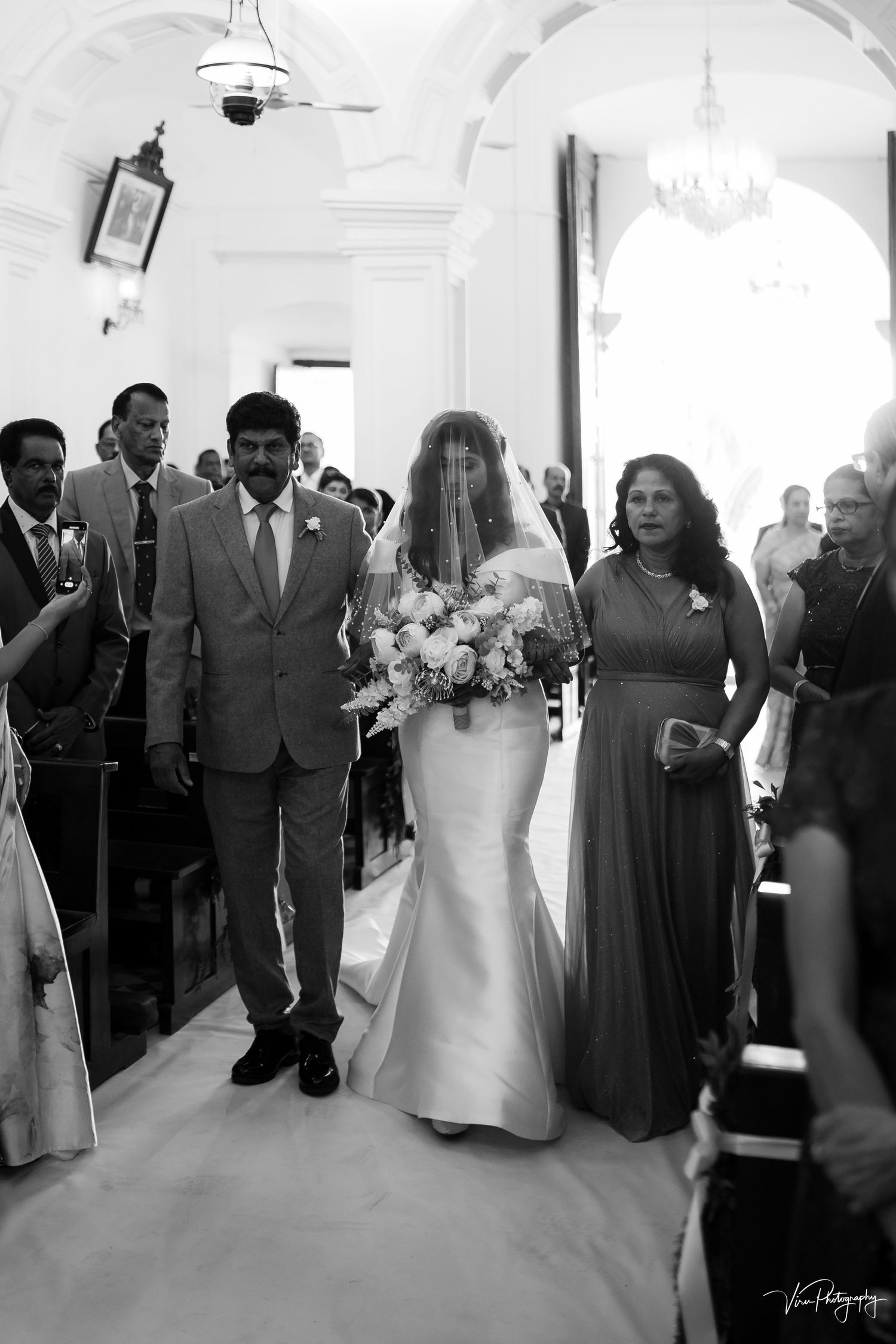 A black and white photo of a wedding processional walking down a brightly lit church aisle.