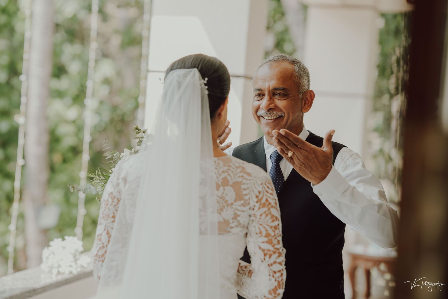 A smiling couple shares an intimate moment during their outdoor wedding ceremony surrounded by white columns and greenery.