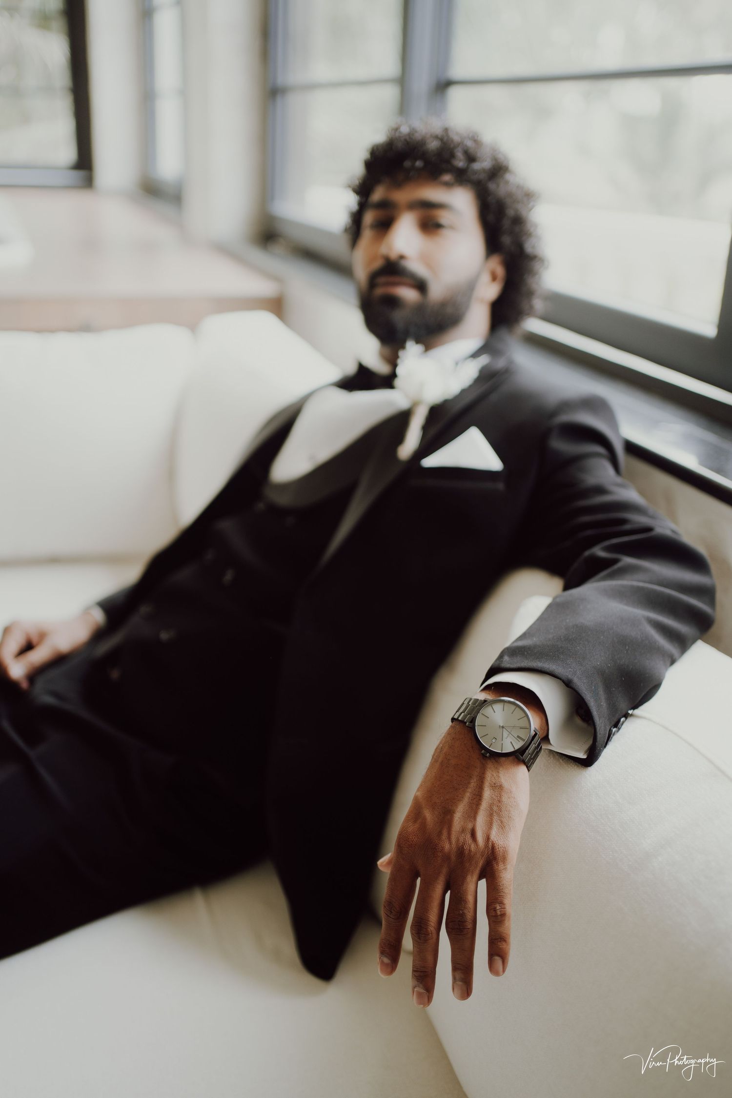 A person in a black tuxedo and bow tie reclines on a white couch near a window, wearing a stylish wristwatch.