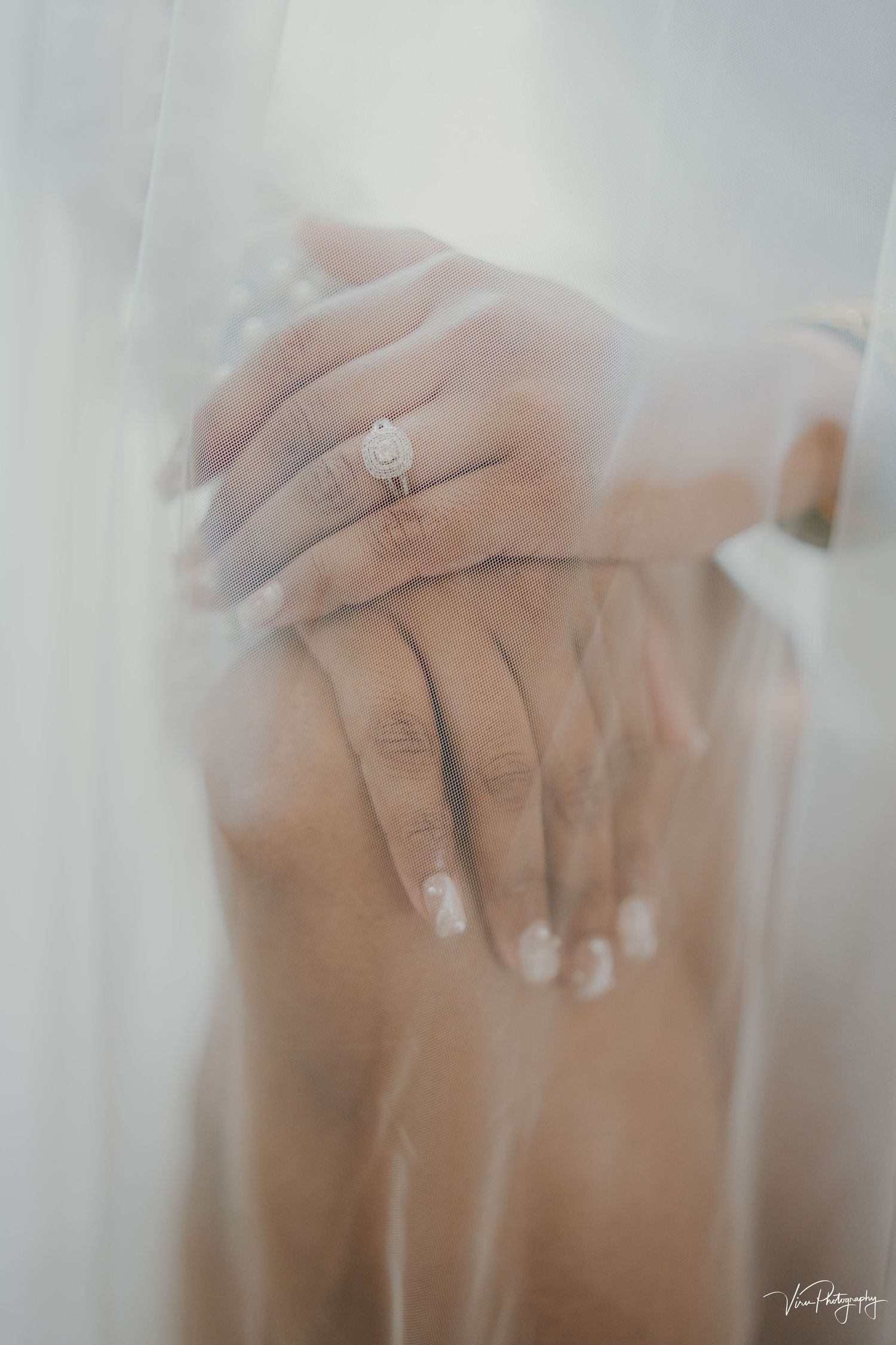 A delicate engagement ring sparkles on a hand with manicured nails, seen through soft white wedding veil fabric.