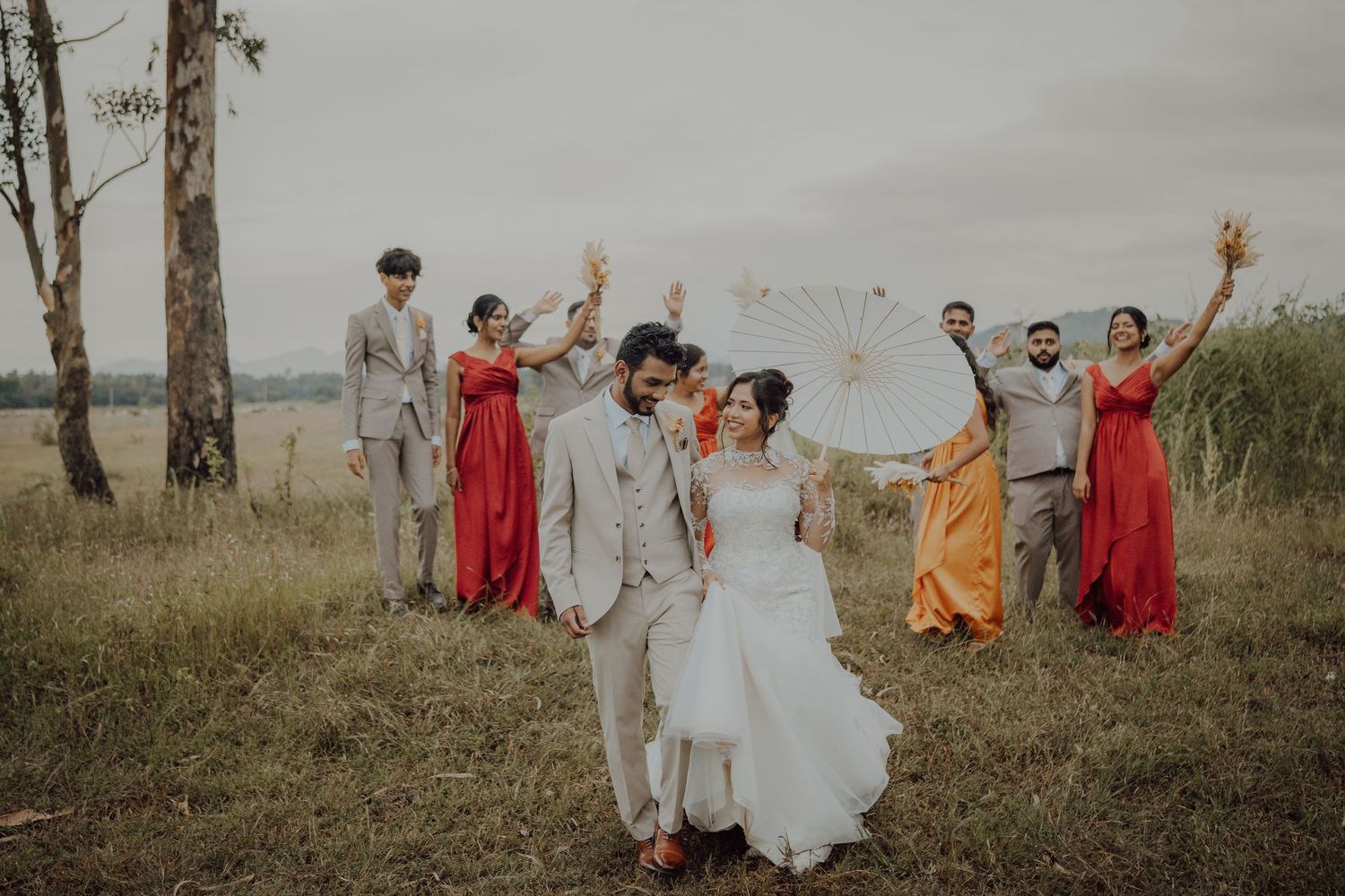Bride and groom walk through a golden field while bridesmaids and groomsmen cheer behind them under a cloudy sky.