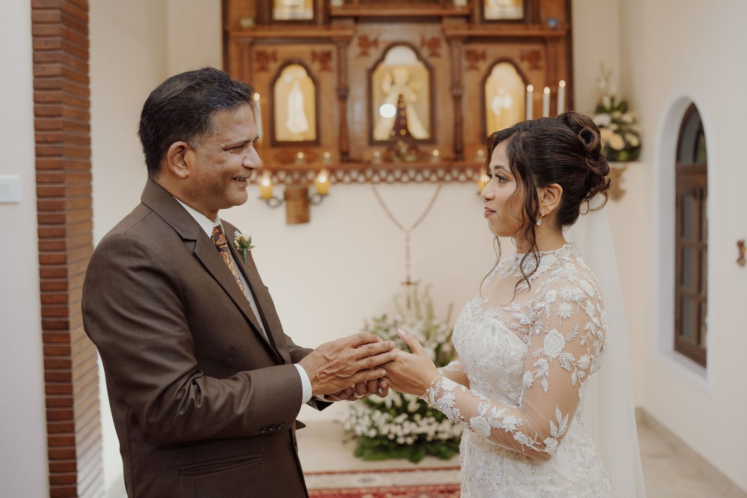 Bride in a lace gown holds hands with an older man in a brown suit inside a church with an ornate altar.