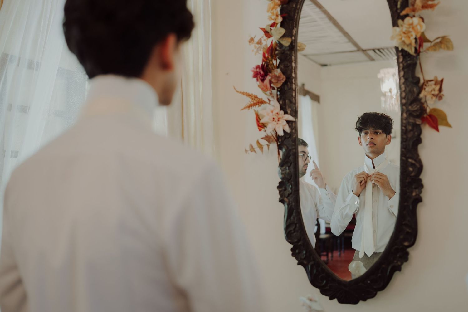 Man in a white shirt adjusts his collar while facing an ornate vintage mirror decorated with dried autumn flowers.