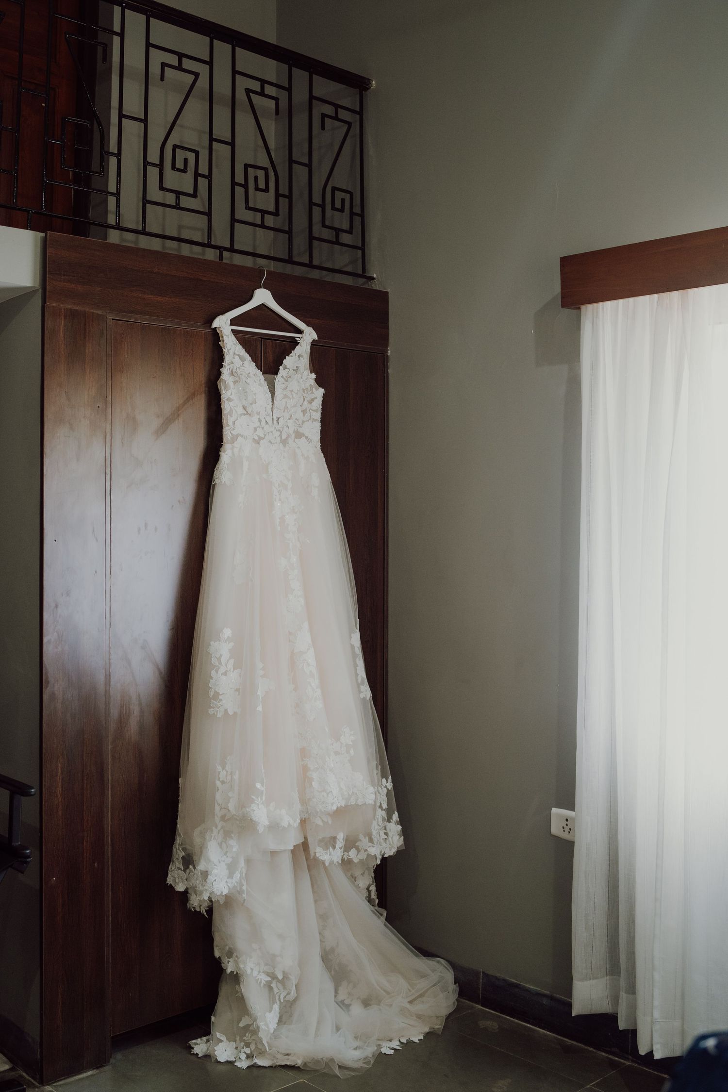 An elegant white lace wedding dress hangs against a dark wooden wardrobe.