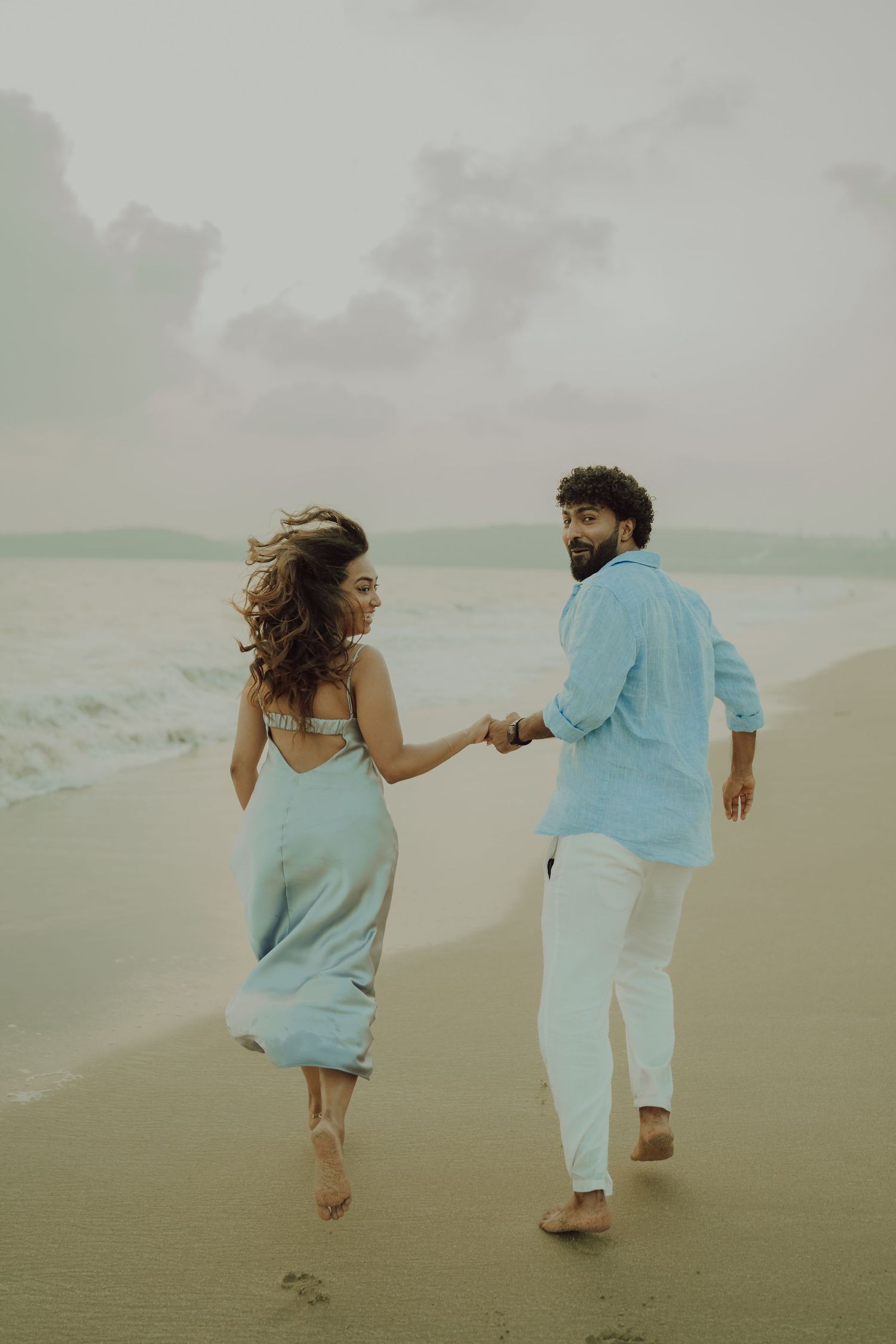A couple holds hands while walking barefoot on a beach at sunset in coordinated light blue and white outfits.