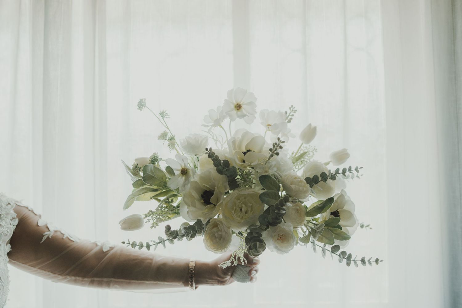 Elegant white floral bouquet being held against a light curtained background.
