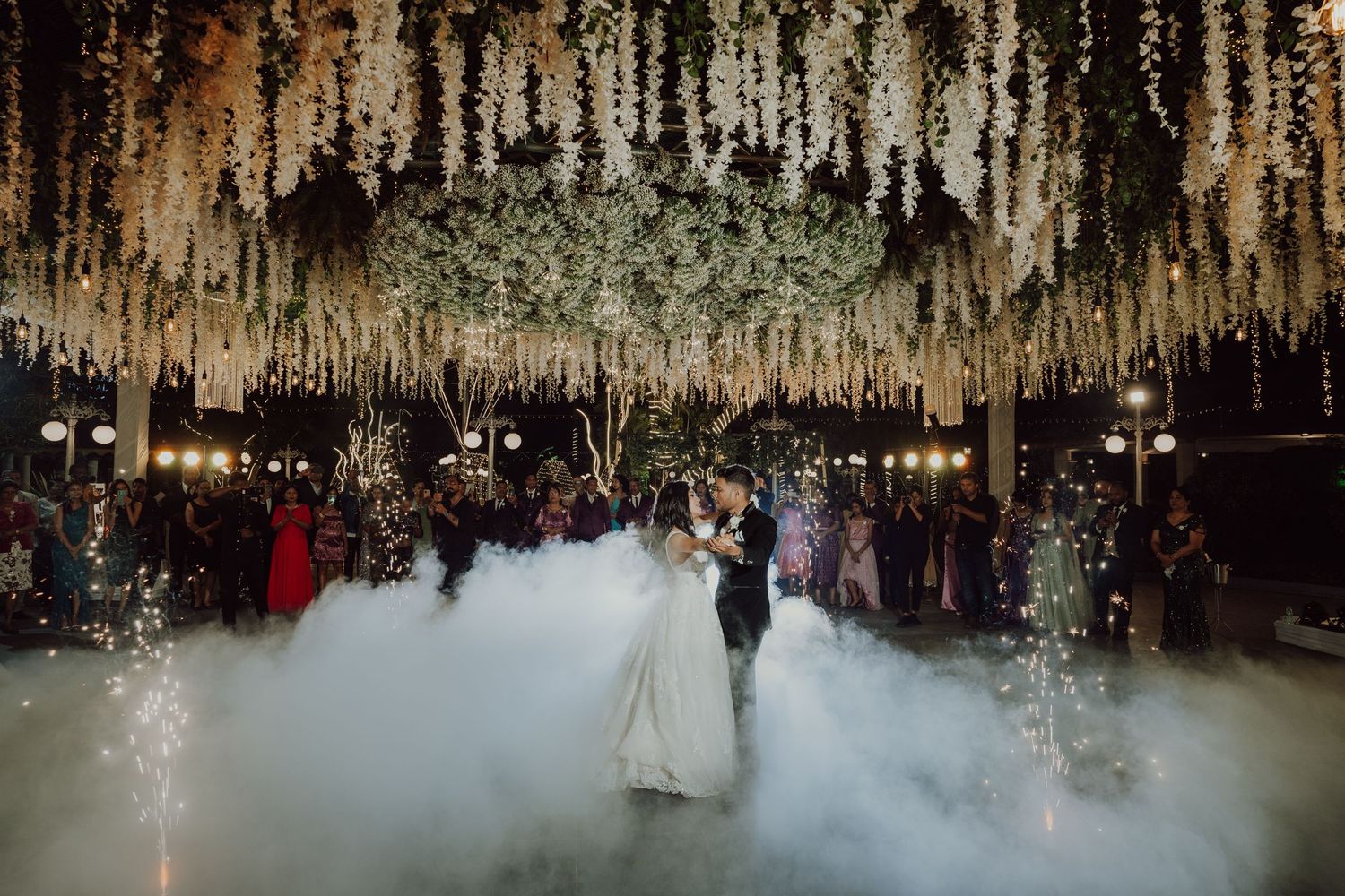 Romantic first dance under a canopy of hanging flowers with atmospheric fog on the dance floor.