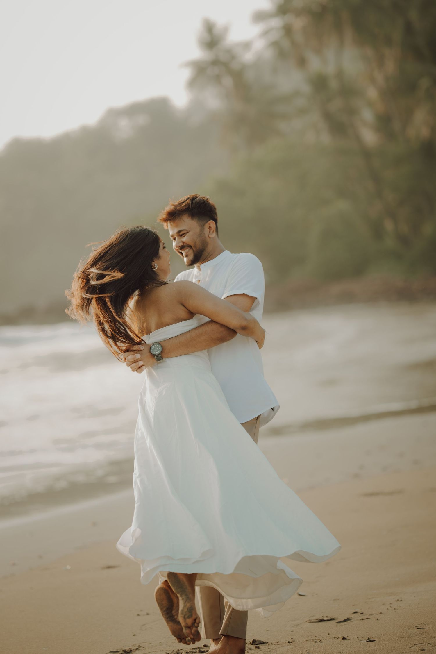 A couple in light flowy clothing dances playfully together on a misty beach at sunset.