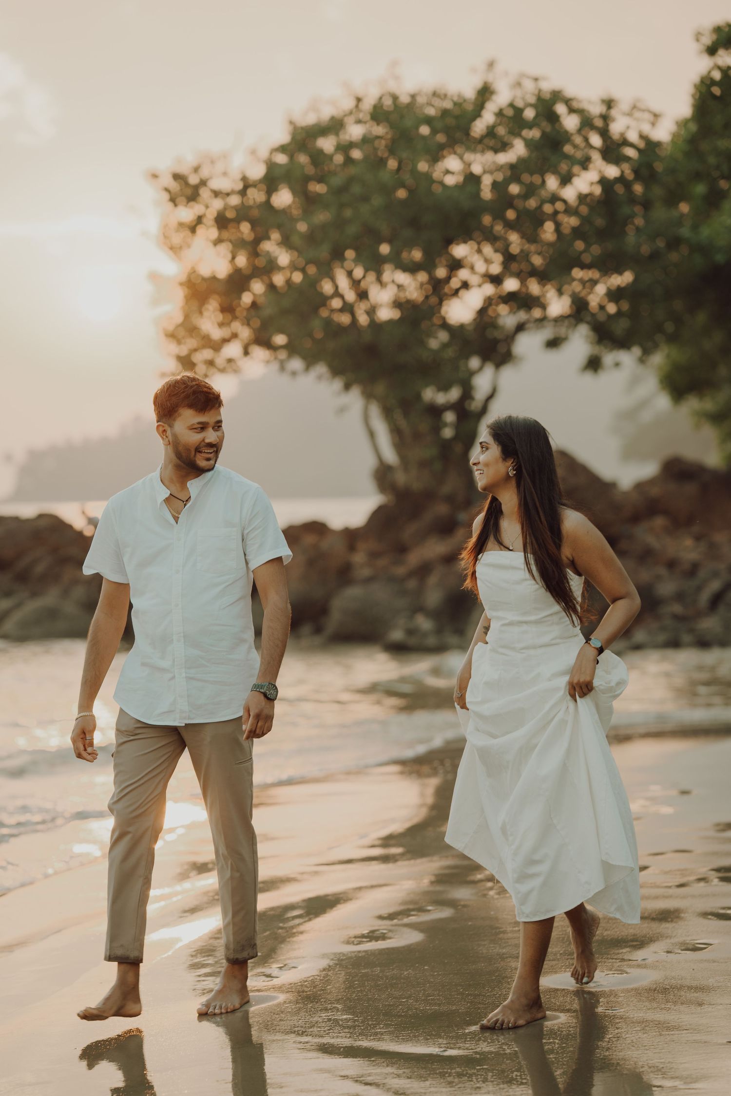 A couple walks playfully along a sandy beach at sunset while wearing light colored casual attire.