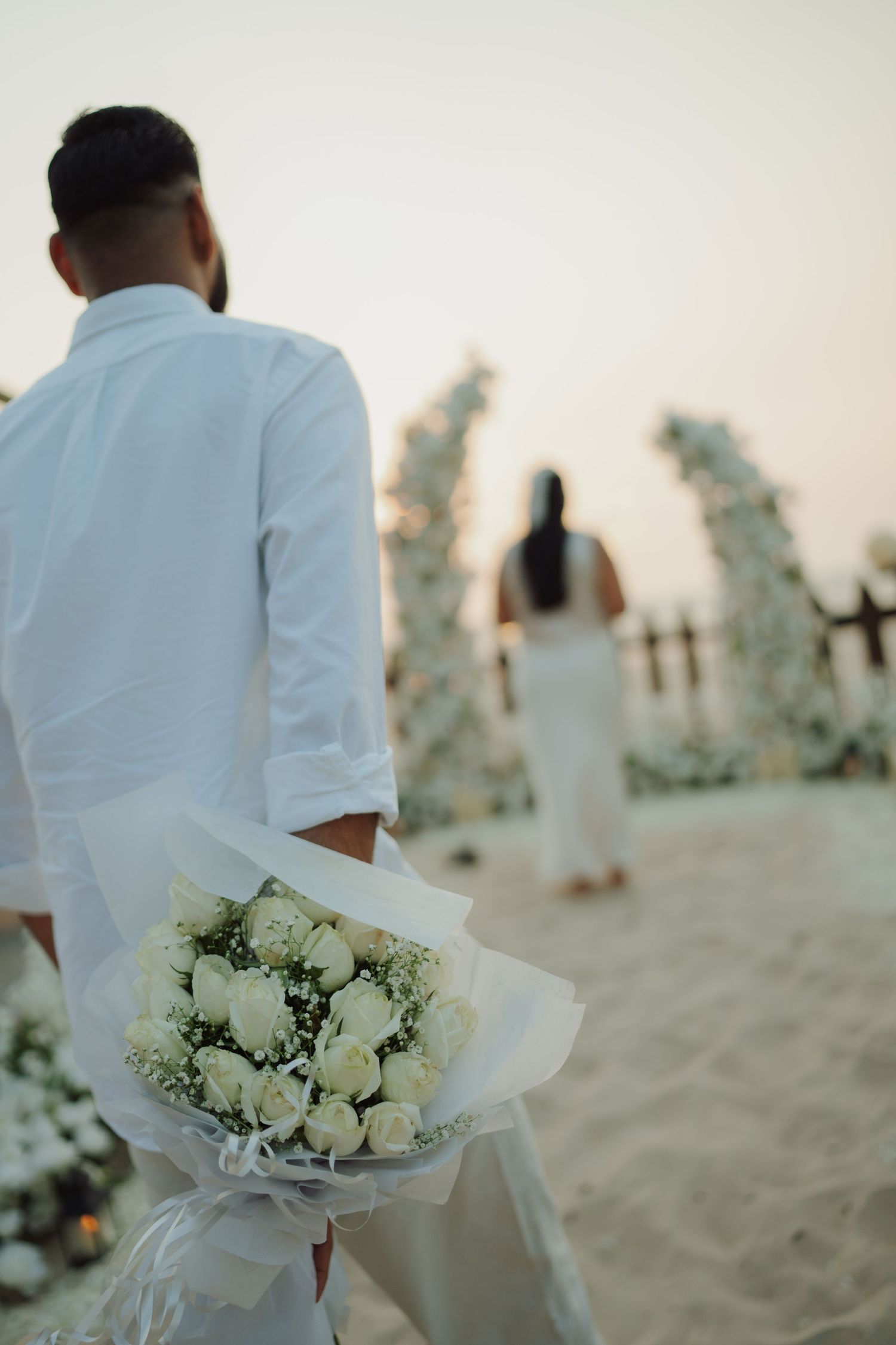 Groom in white shirt holds bouquet of white roses at beach wedding ceremony with floral arch decorations at sunset.