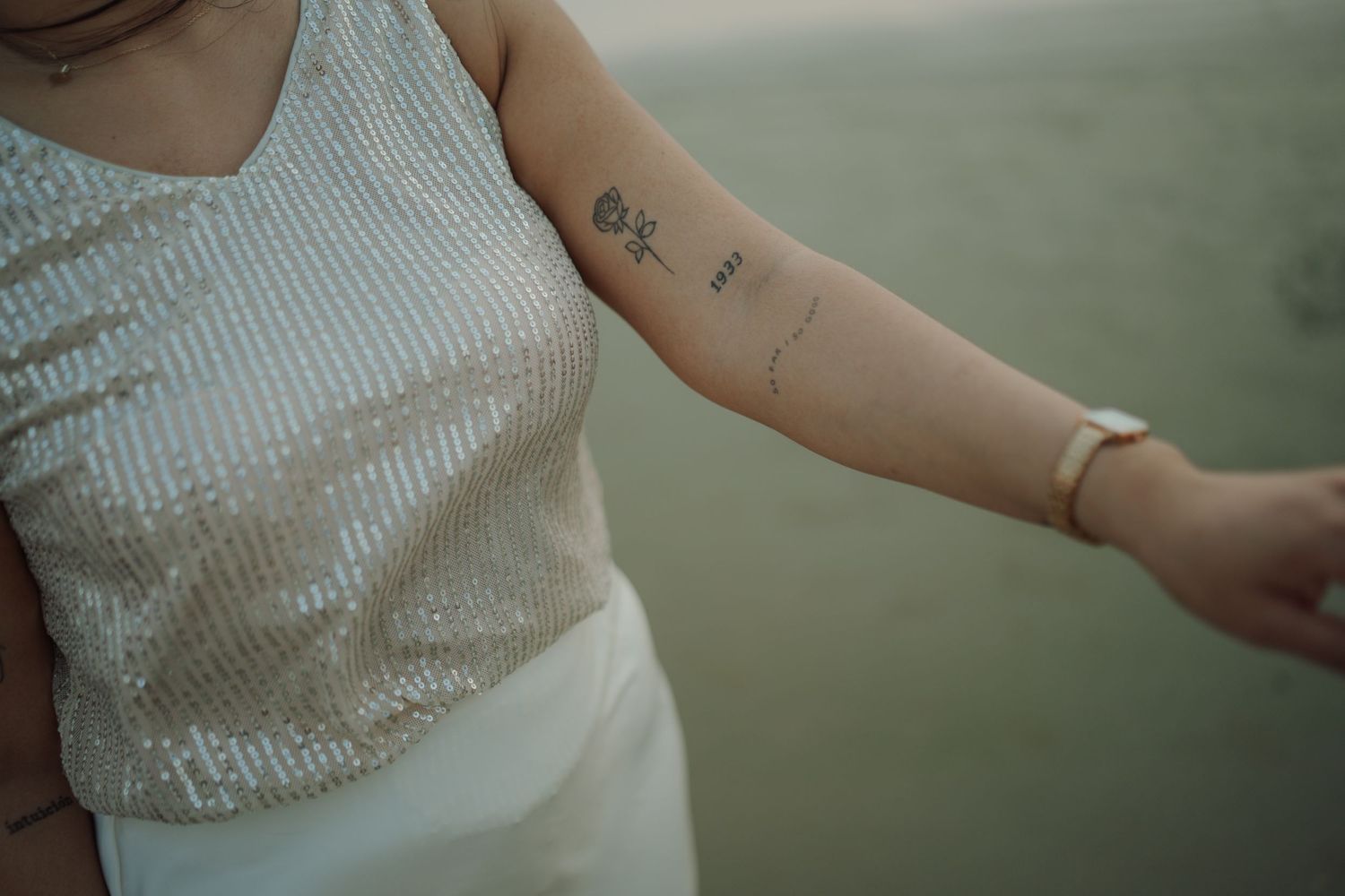 Woman in sequin top with floral arm tattoo and bracelet holding hands outdoors.