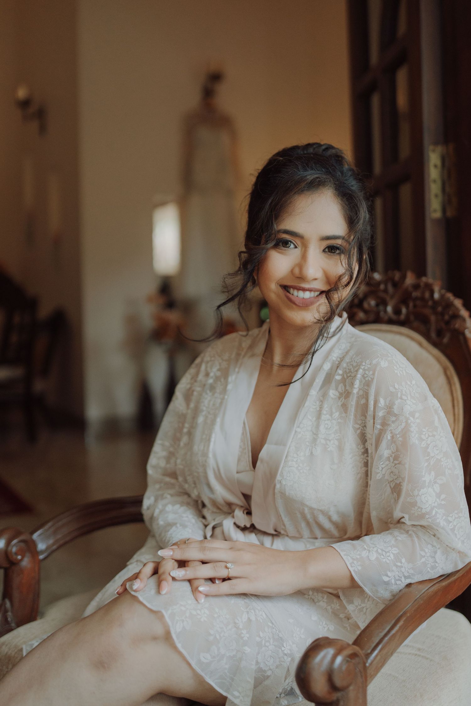 Smiling bride in a lace robe with her hair in an elegant updo sits in a carved wooden chair while getting ready for her wedding.