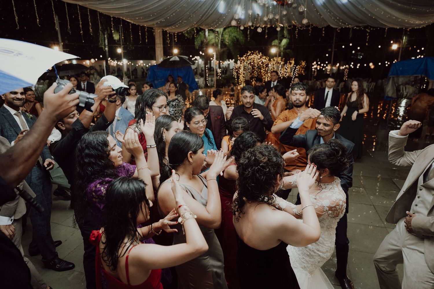 Wedding guests dance and clap enthusiastically at a lively outdoor reception lit by warm string lights at night.