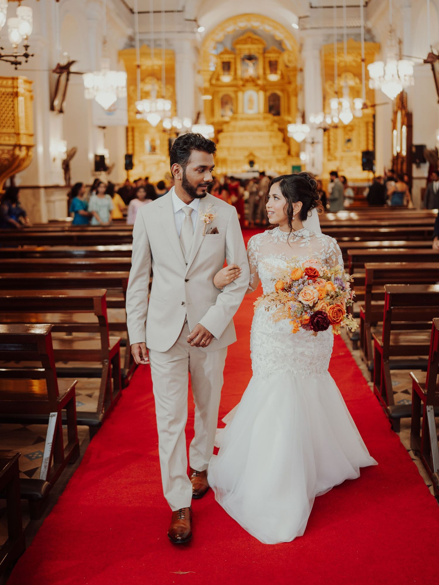 Bride and groom walk arm in arm down a red carpet aisle in a church lined with wedding guests.