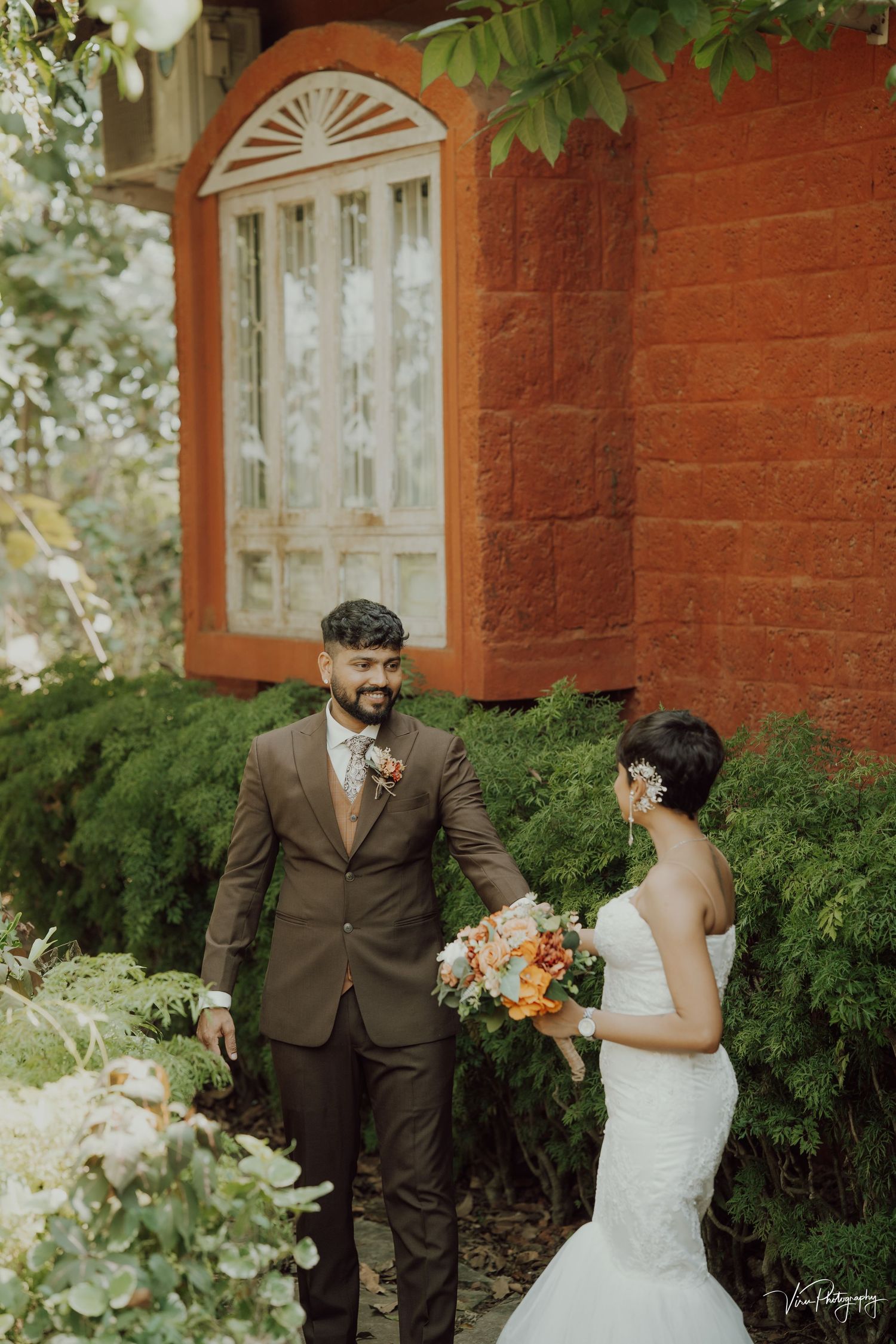 A wedding couple stands in front of a rustic red brick wall with an elegant arched window and lush greenery.