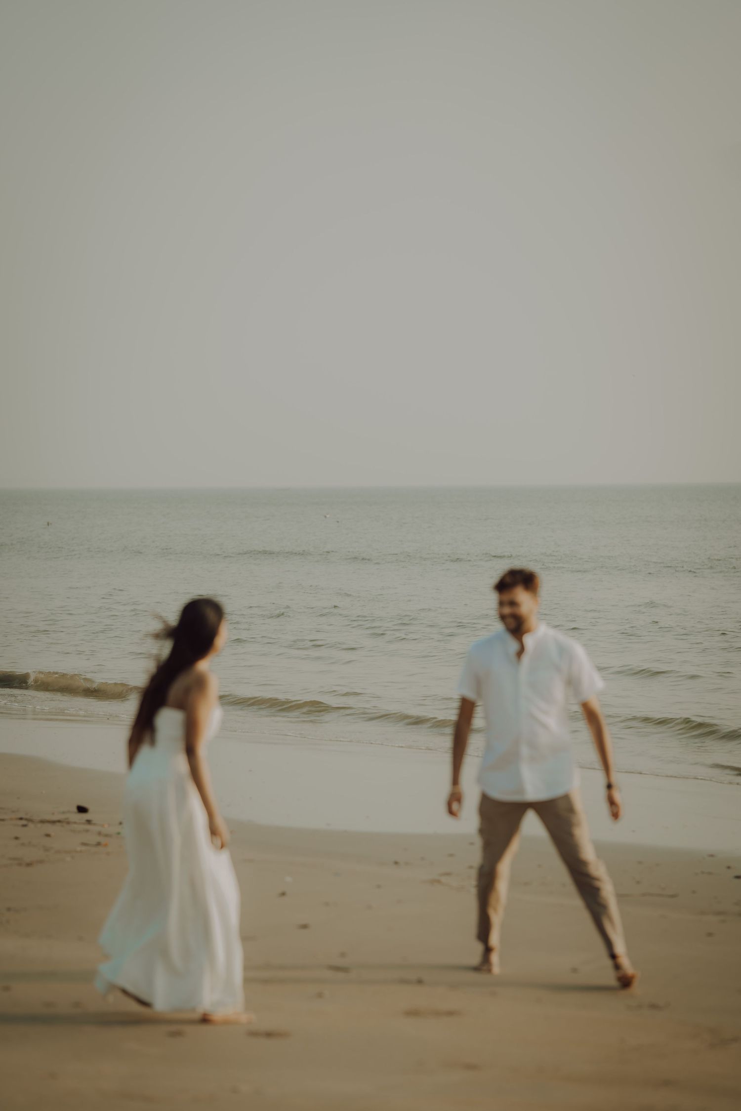 A playful couple runs and dances together on a serene beach during an overcast day.