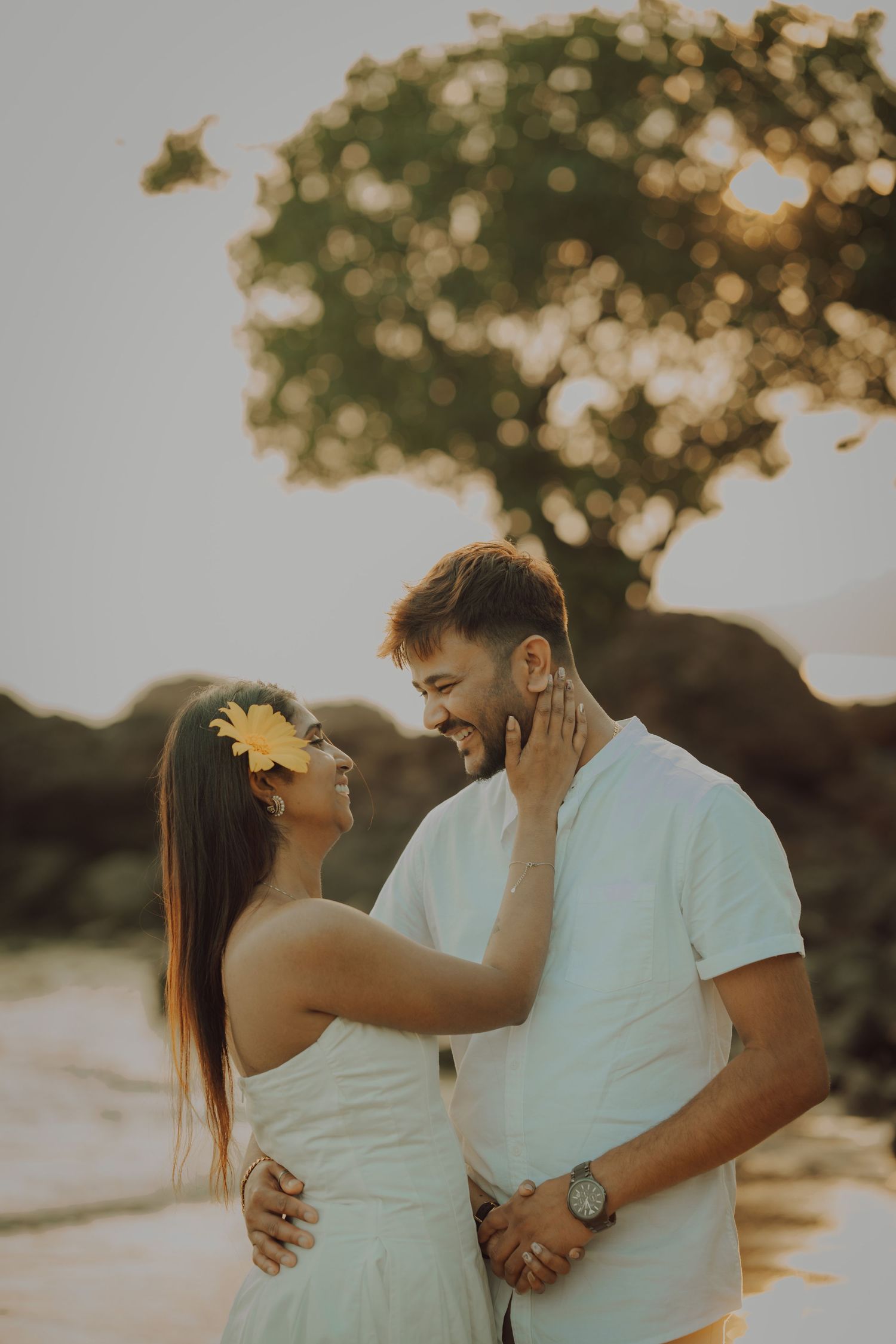 A romantic couple in white outfits share an intimate moment at sunset with bokeh lights from trees in the background.