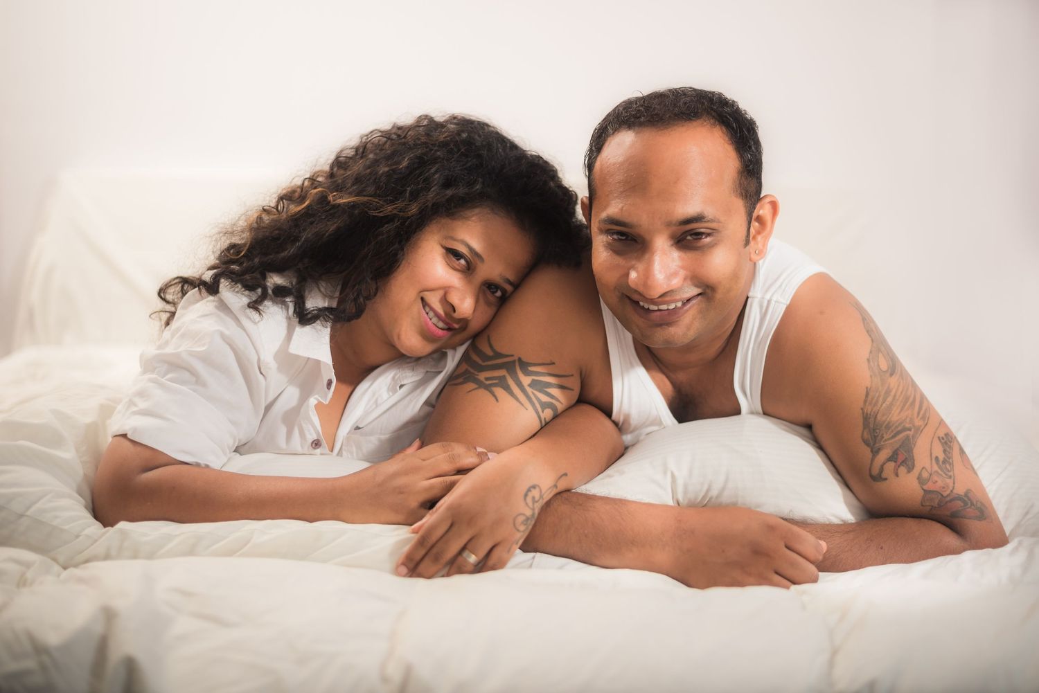 A couple lies together on white bedding while sharing an intimate moment together.