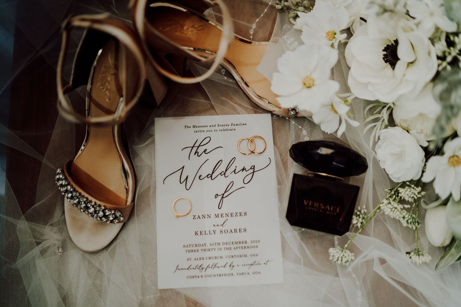 Wedding details flatlay with invitation, jeweled bridal shoes, perfume bottle and white floral arrangements on wooden surface.