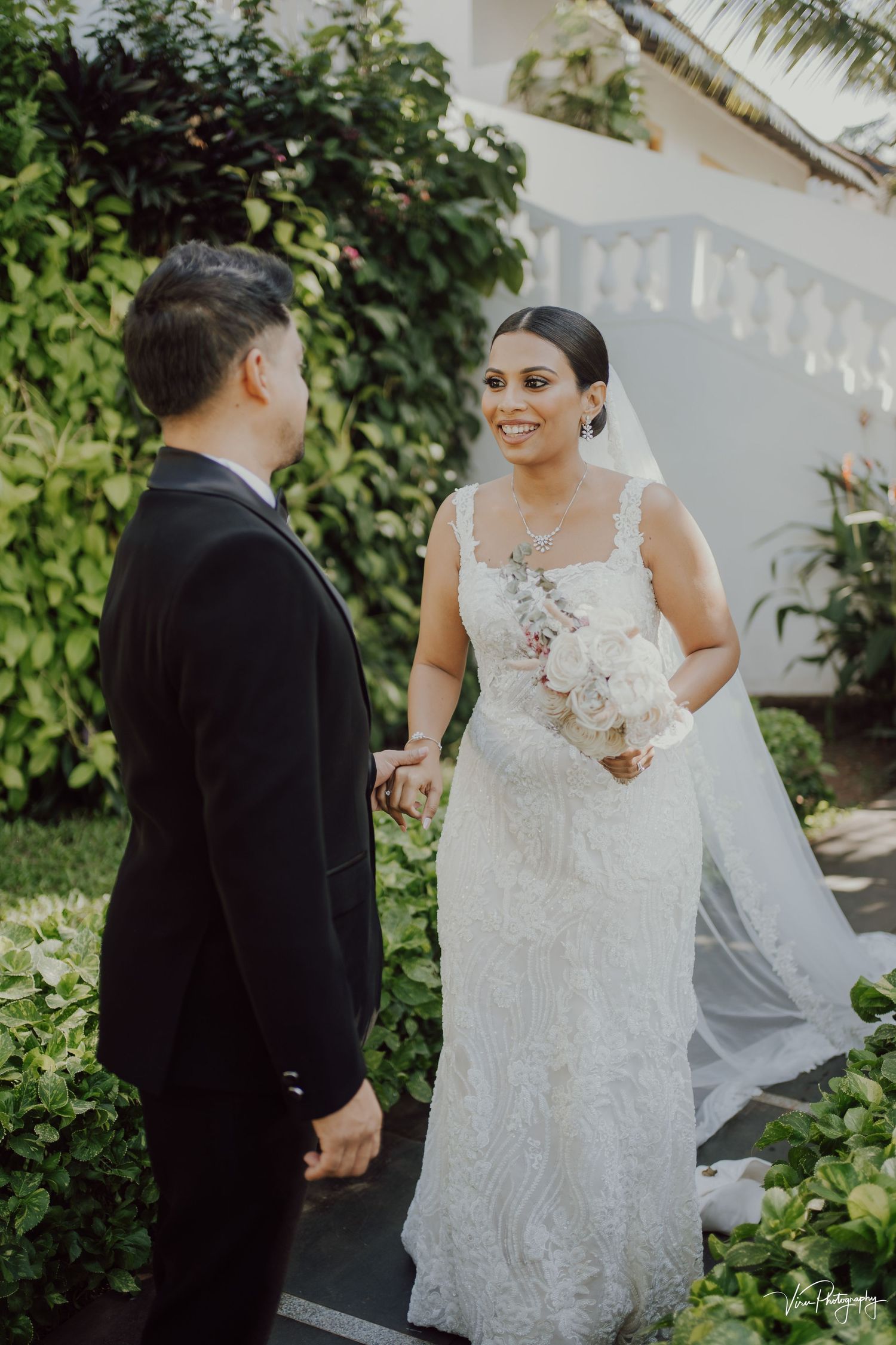 A couple shares a joyful wedding moment in a garden setting with the bride wearing a detailed lace gown and flowing veil.