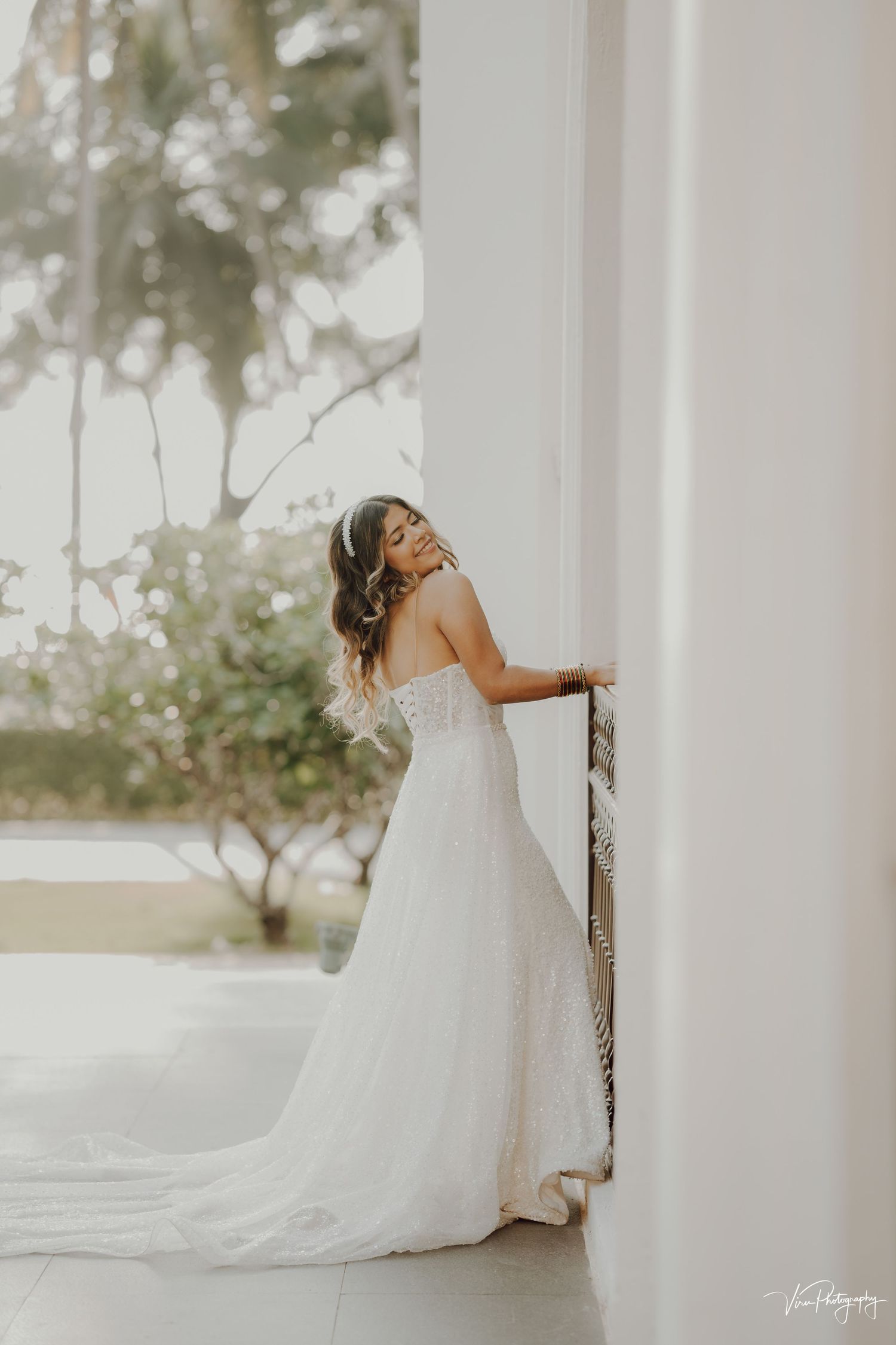 A bride in a flowing white strapless wedding gown with train leans against a white pillar in romantic outdoor setting.