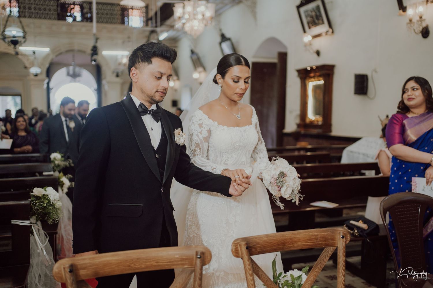 A couple stands together during a traditional church wedding ceremony with wooden pews decorated with floral arrangements.