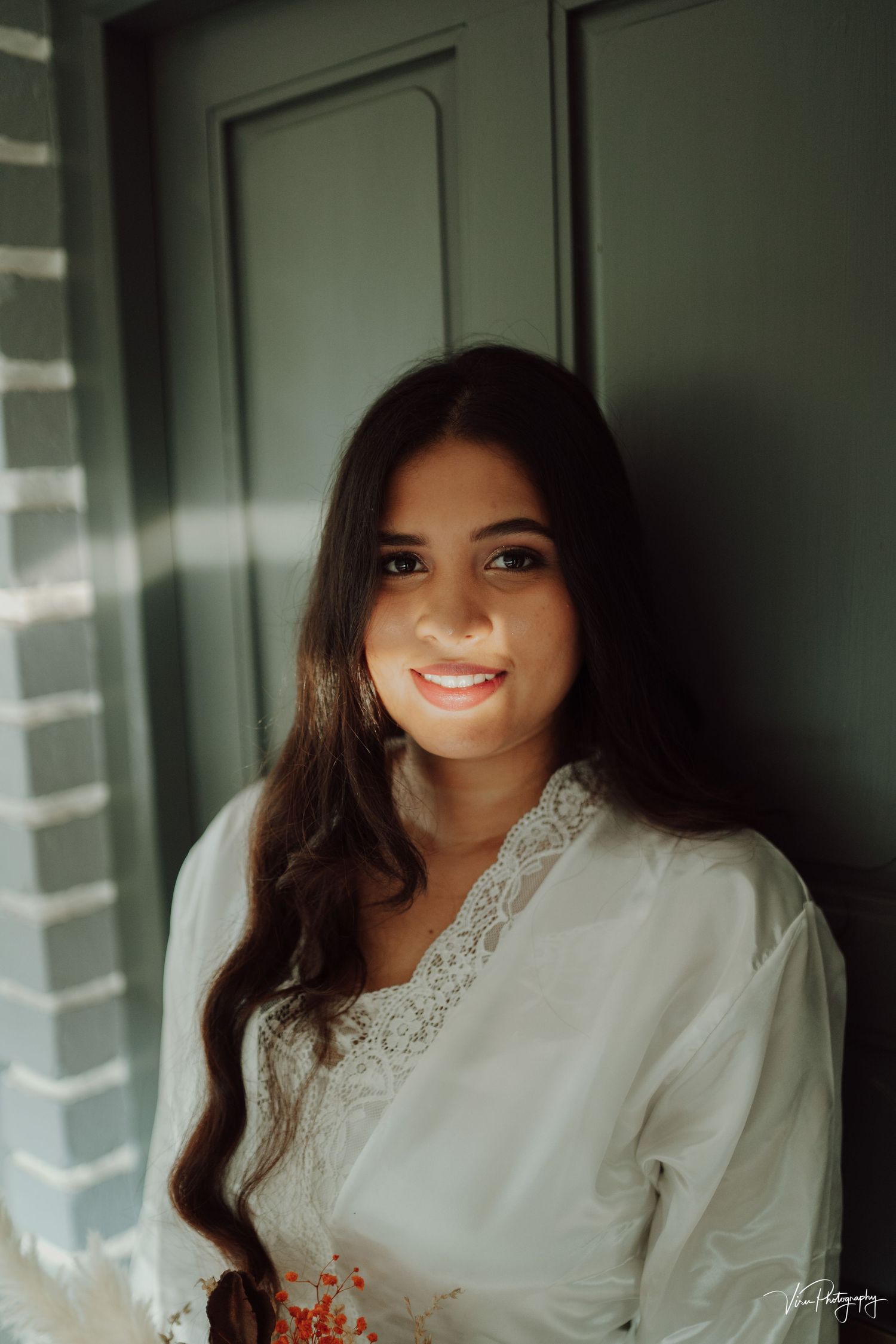 A person in a white lace-trimmed top smiles while standing near a window with natural lighting.