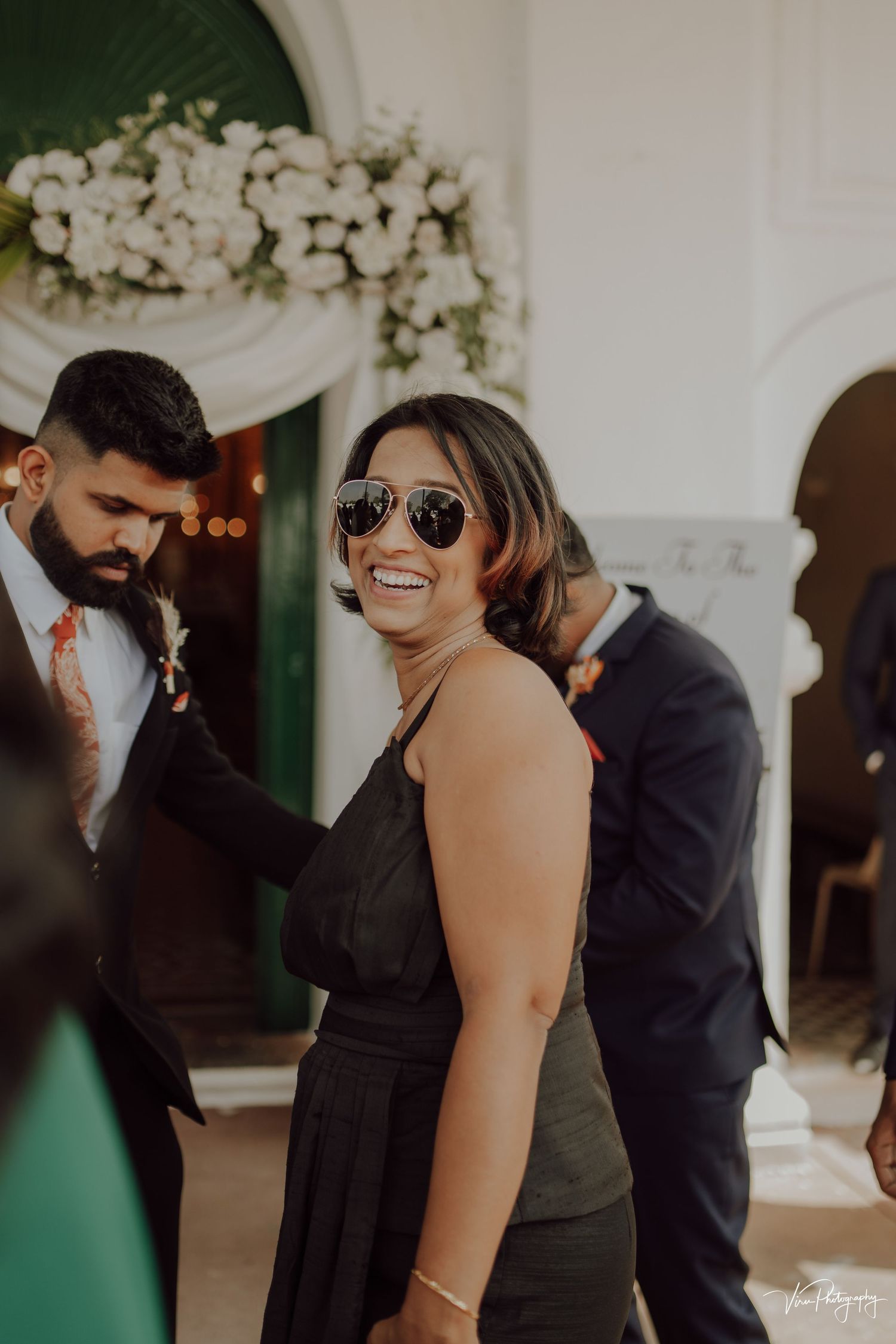 Wedding guest in a black dress and sunglasses smiles joyfully at an elegant reception with white floral decorations.