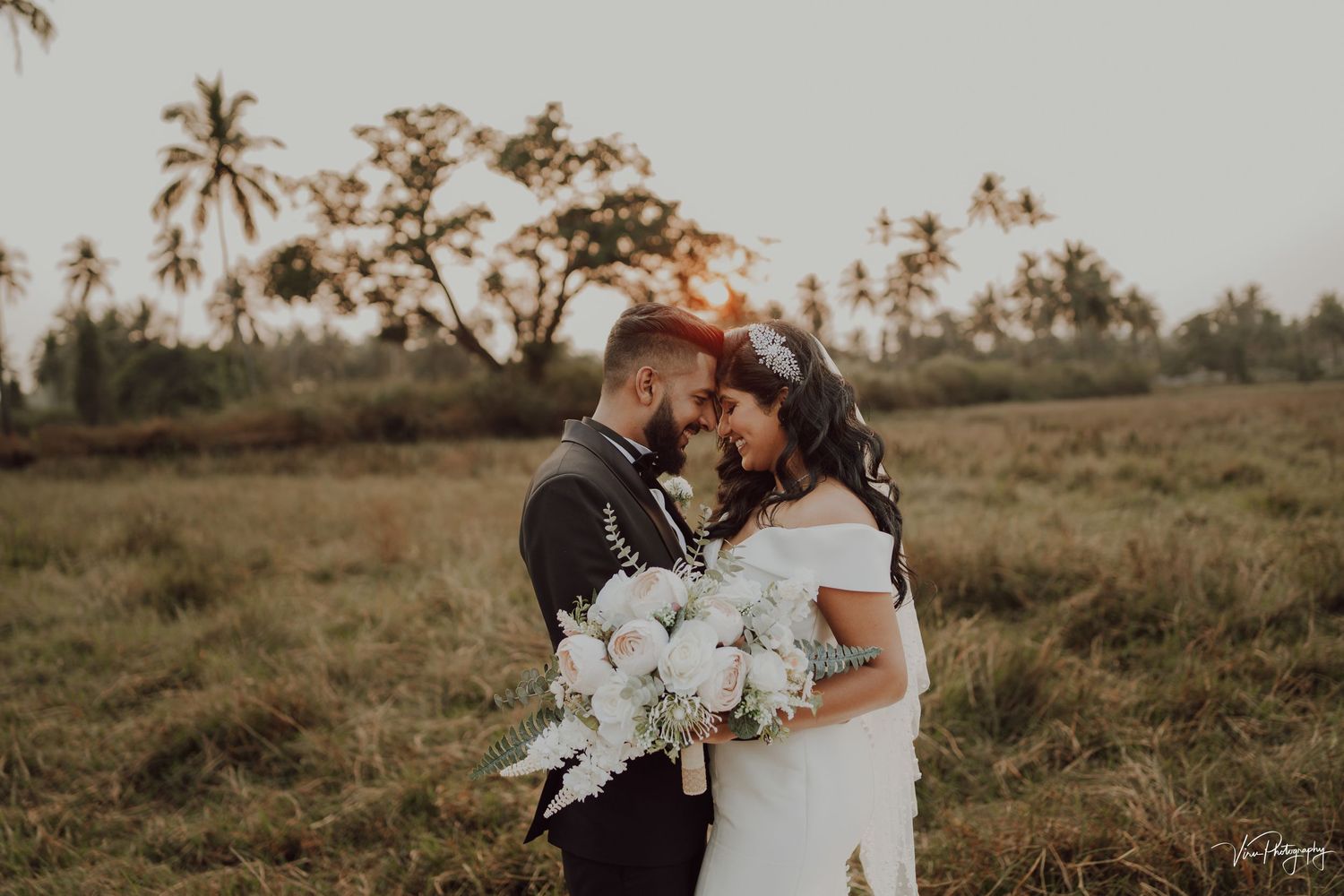 A romantic couple embraces in a field at sunset with palm trees and a white floral bouquet.