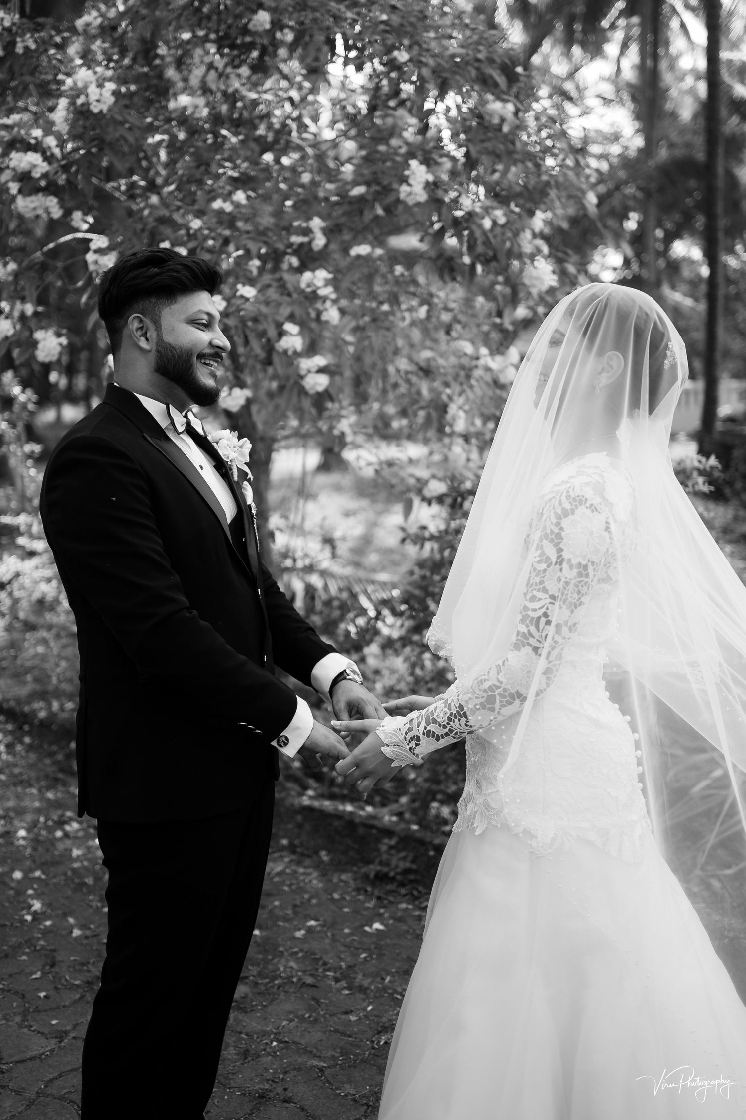 A black and white wedding photo capturing a couple holding hands in a garden setting with a long flowy veil.