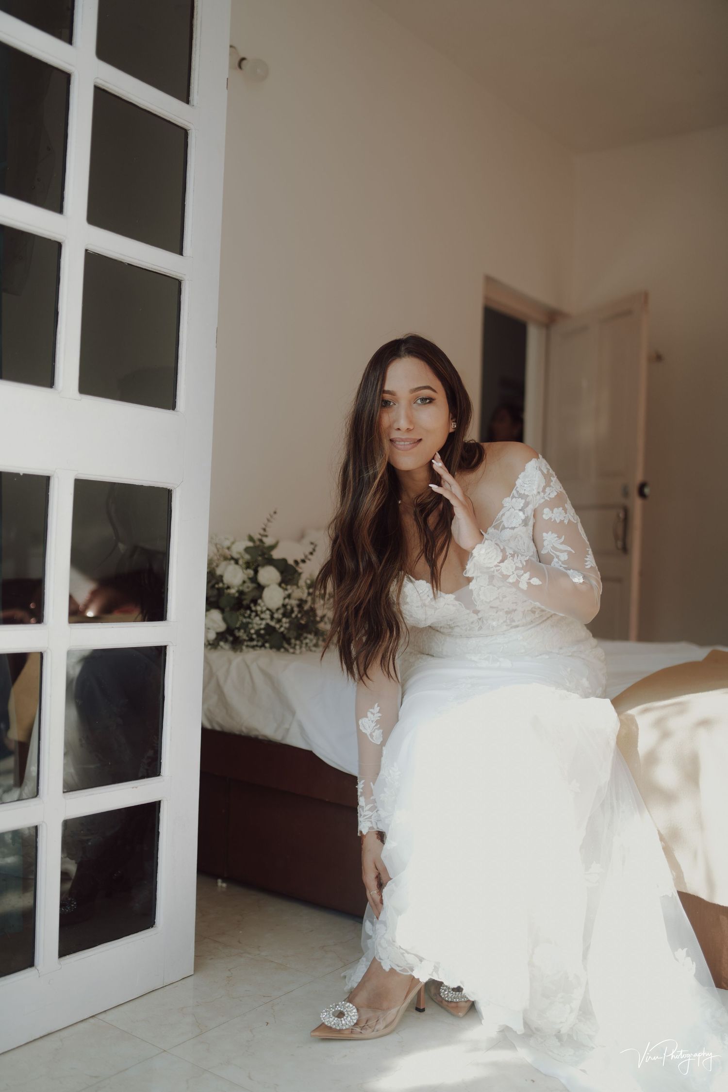 Bride in an elegant white lace wedding dress with long sleeves adjusts her shoe in a bright interior space.