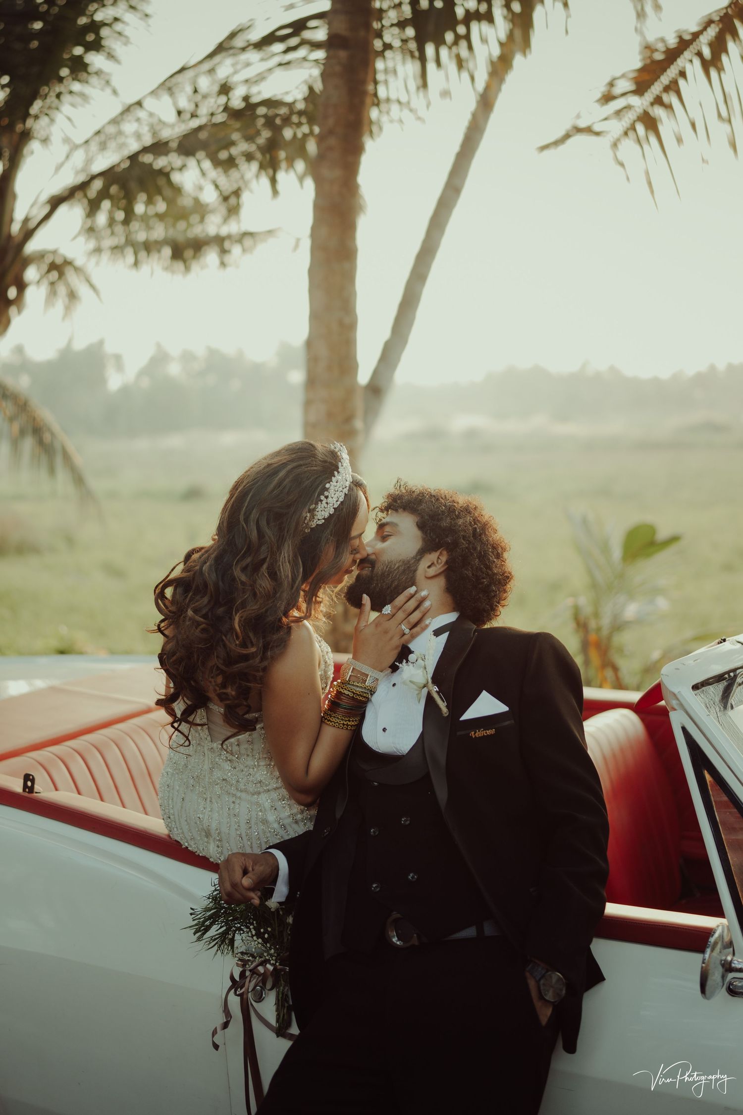 A happy couple shares a romantic moment beside a vintage red car with palm trees in the background at sunset.