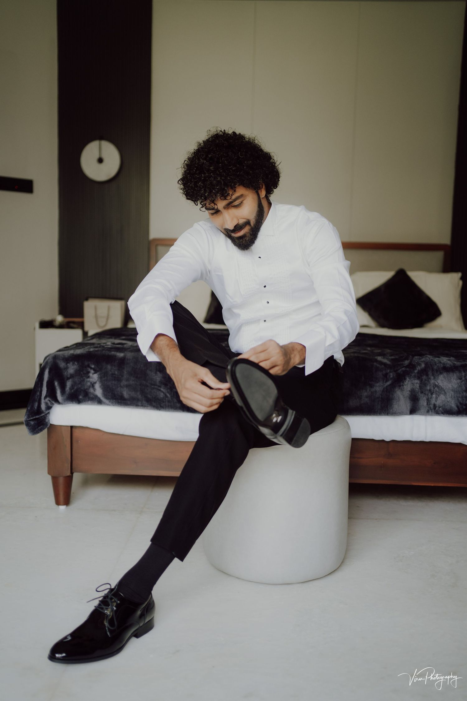 A person in a white shirt and black pants sits on a white ottoman, adjusting their formal black dress shoes.